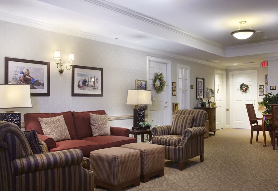 Comfortable senior living common room with a sofa, striped armchairs, ottomans, lamps, framed wall art and a dining area in the background.