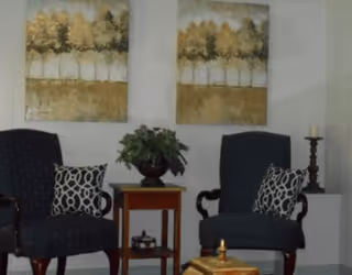 Two upholstered armchairs with patterned pillows flank a small table holding a potted plant beneath two tree paintings, with a candle on a pedestal and a decorative ottoman in front.