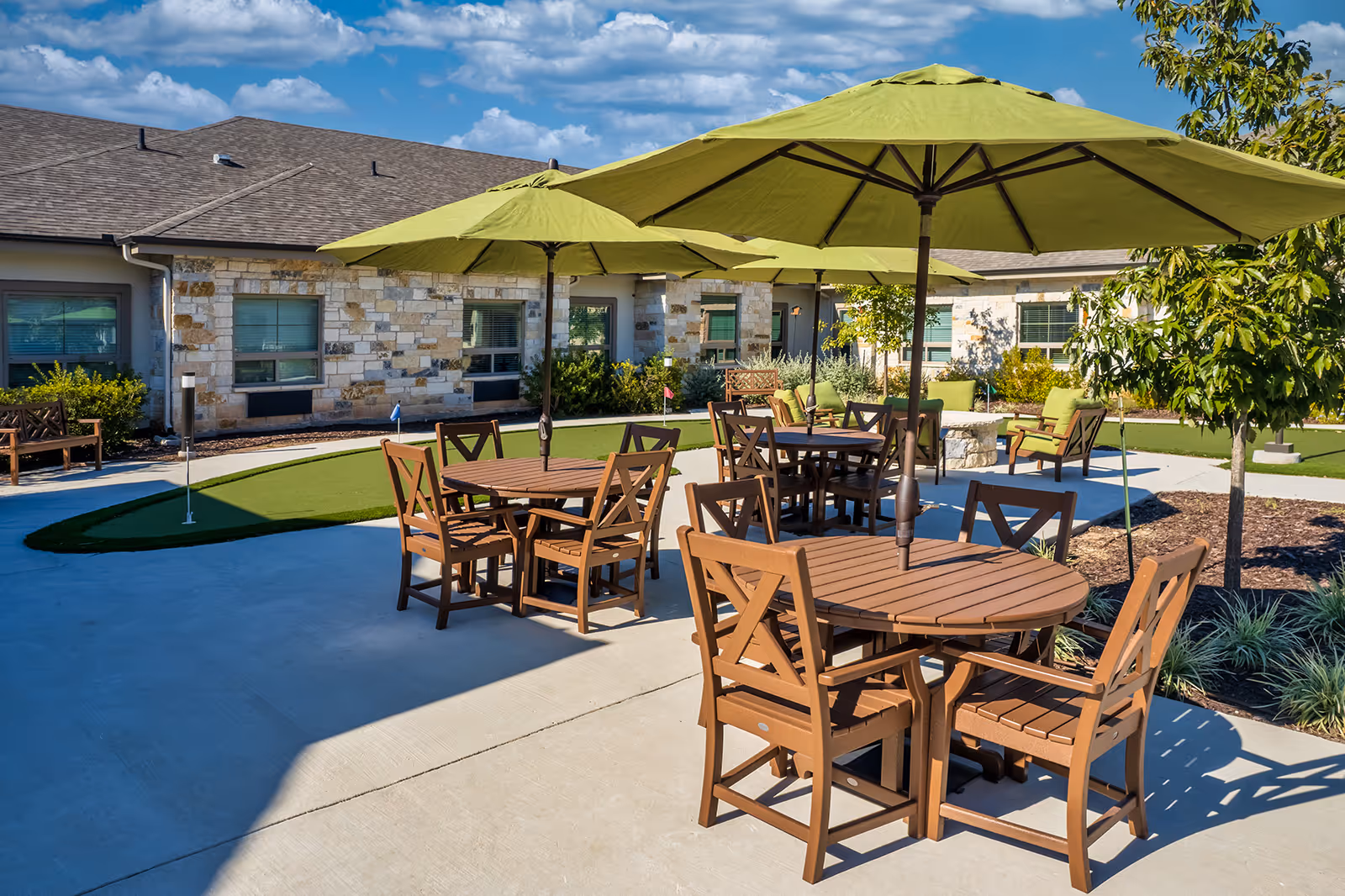 Outdoor courtyard with round wooden tables and green umbrellas on a concrete patio and a small putting green in front of a low stone building.