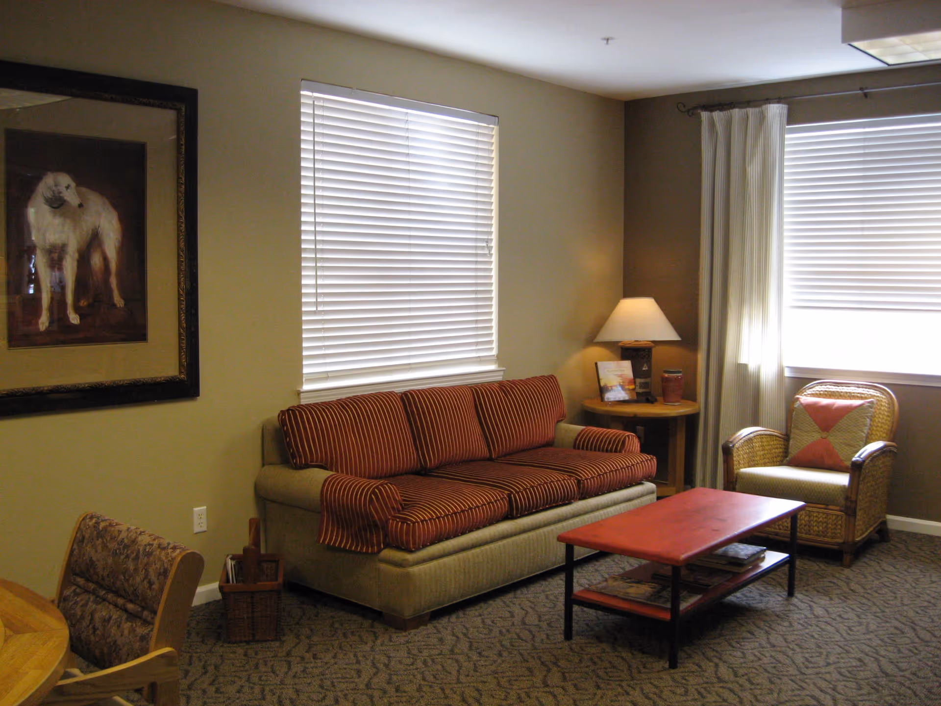 A cozy living room with a beige sofa featuring red striped cushions, a wooden coffee table with magazines underneath, a wicker armchair with a decorative pillow, a round wooden side table with a lamp and some items on it, two windows with white blinds, and a framed painting of a dog on the wall.