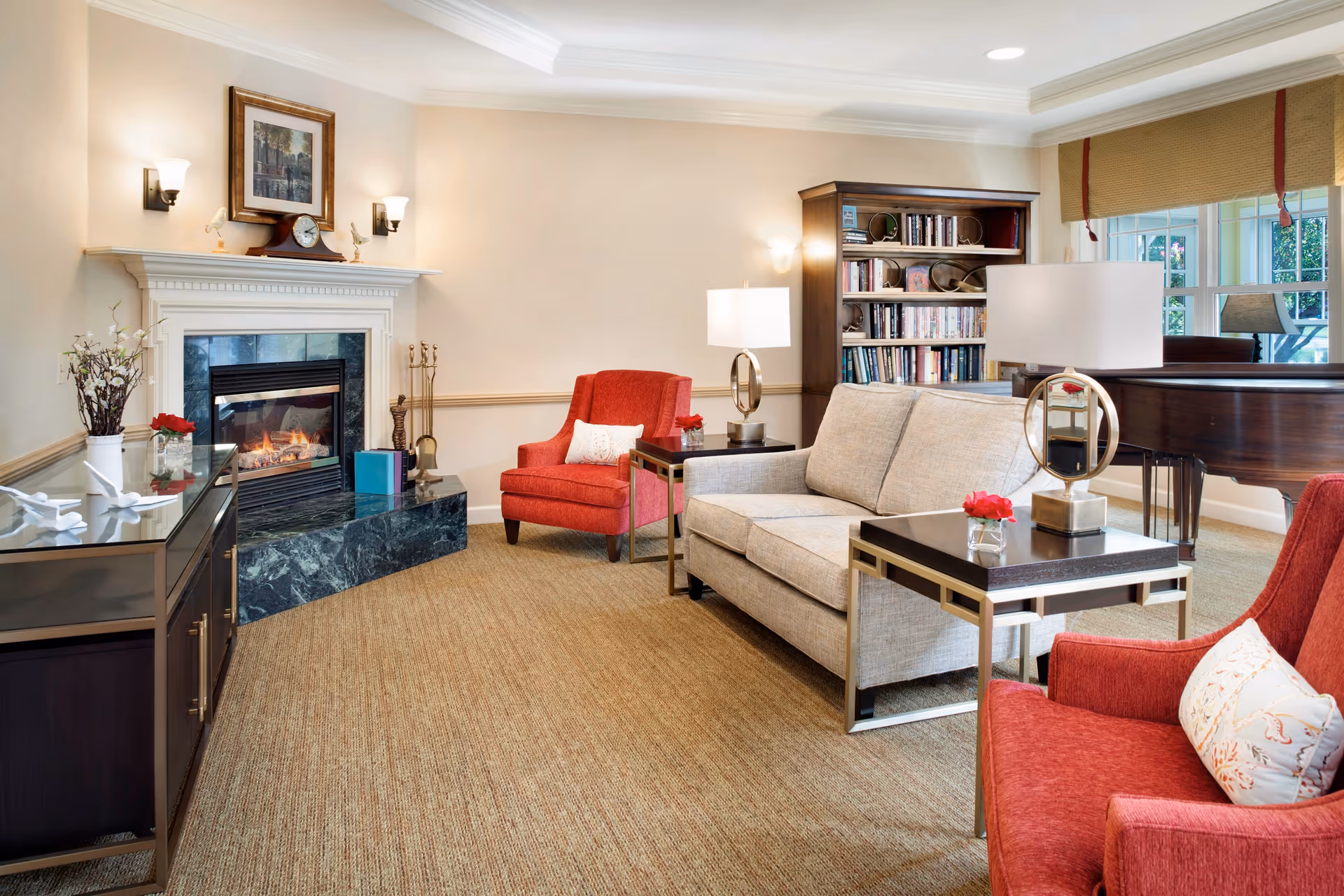 Bright common room with a fireplace, sofa, red armchairs, bookshelves and a piano.