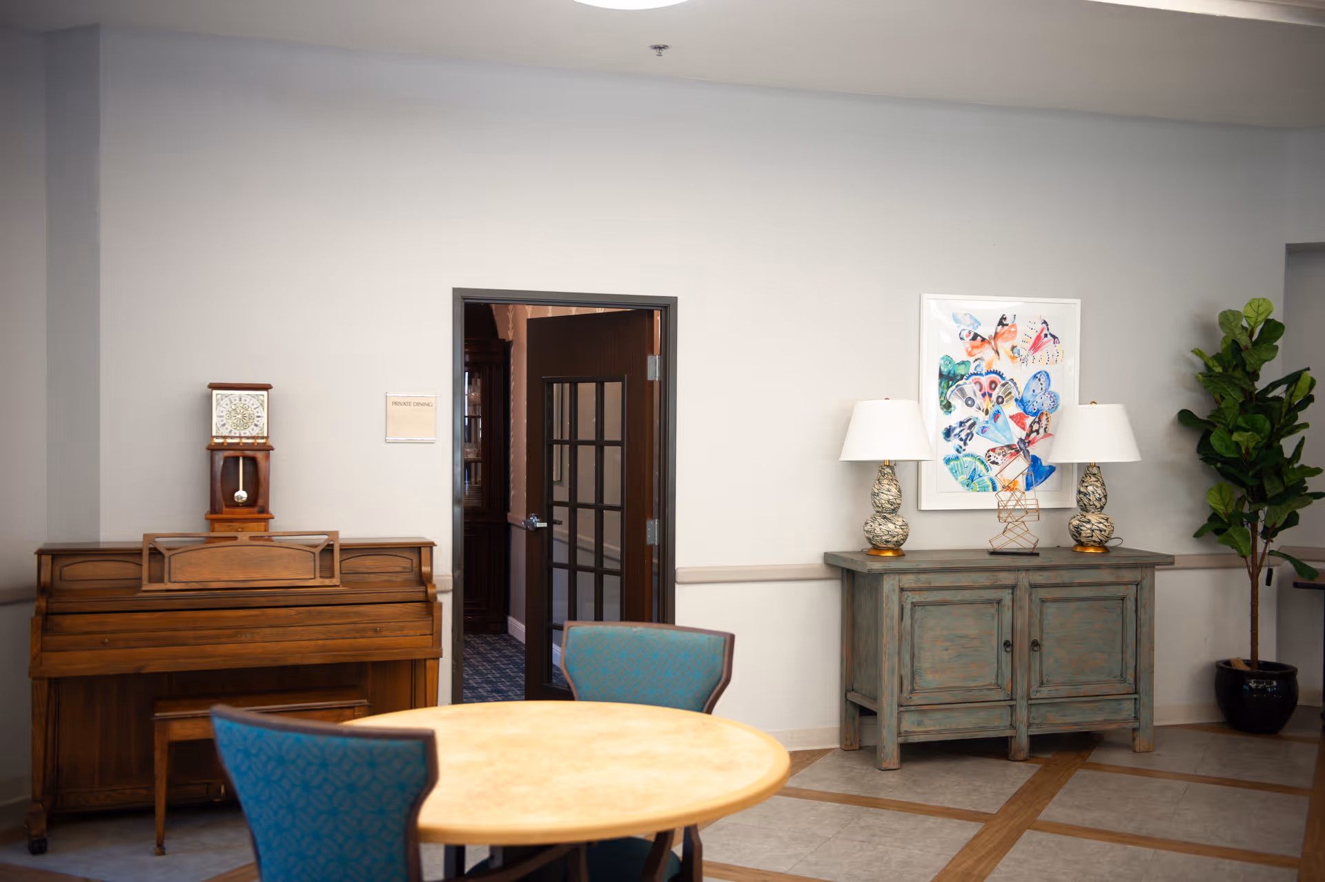 A room with a round wooden table and blue cushioned chairs in the foreground. Against the wall, there is a wooden piano with a clock on top and a sign next to a door labeled 'Private Dining.' On the right side, there is a rustic wooden cabinet with two decorative lamps and a colorful butterfly-themed artwork above it. A tall green potted plant is placed in the corner.