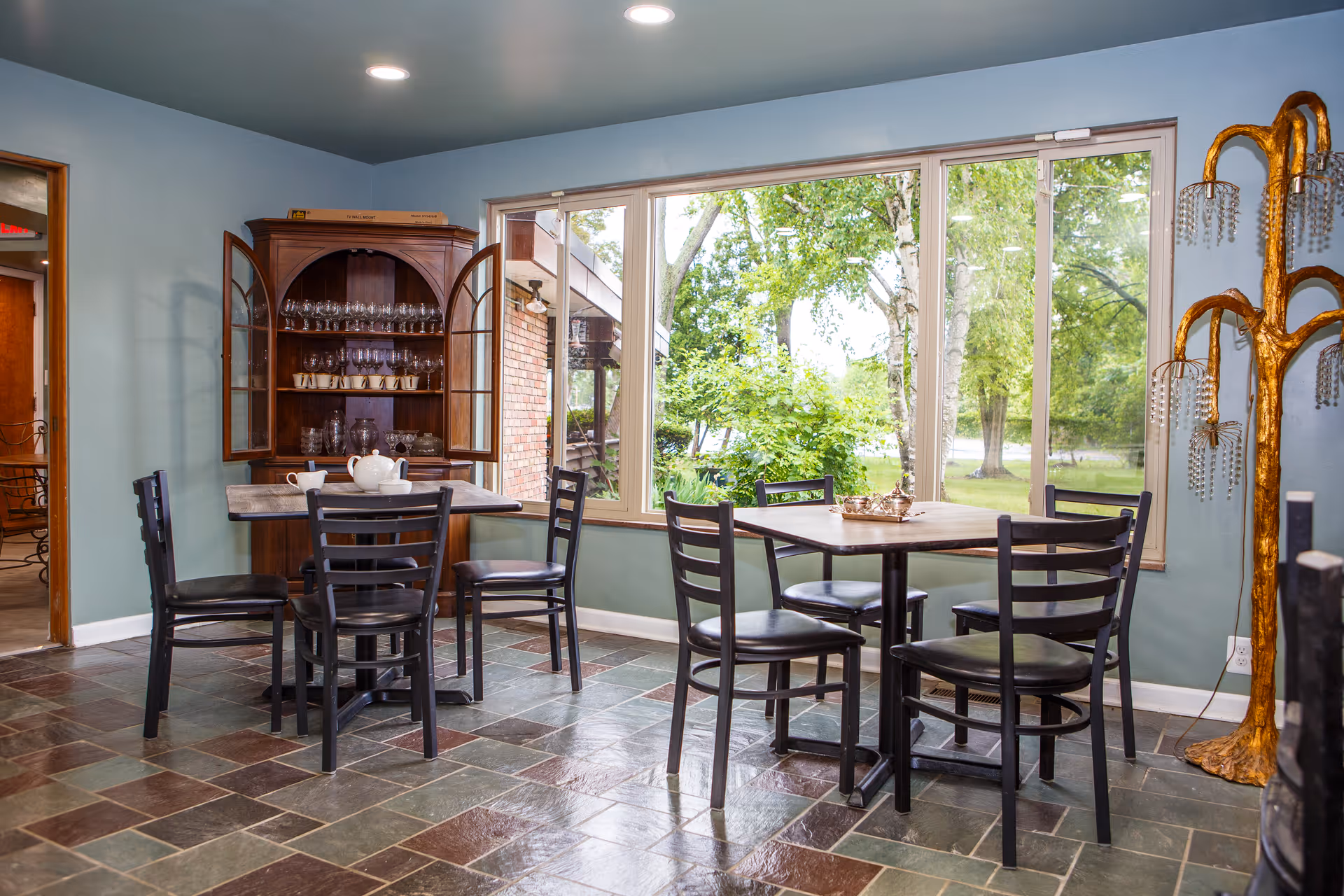 Dining area with two wooden tables and black chairs next to large windows overlooking a green yard.