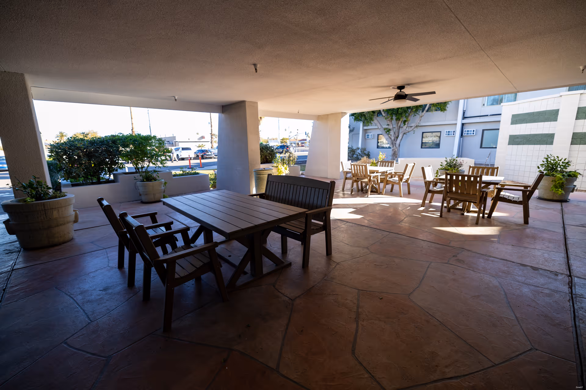 Covered outdoor patio area with multiple wooden tables and chairs arranged for seating. Large potted plants are placed around the space, and a ceiling fan is mounted overhead. The area is adjacent to a building with windows and some greenery visible in the background.