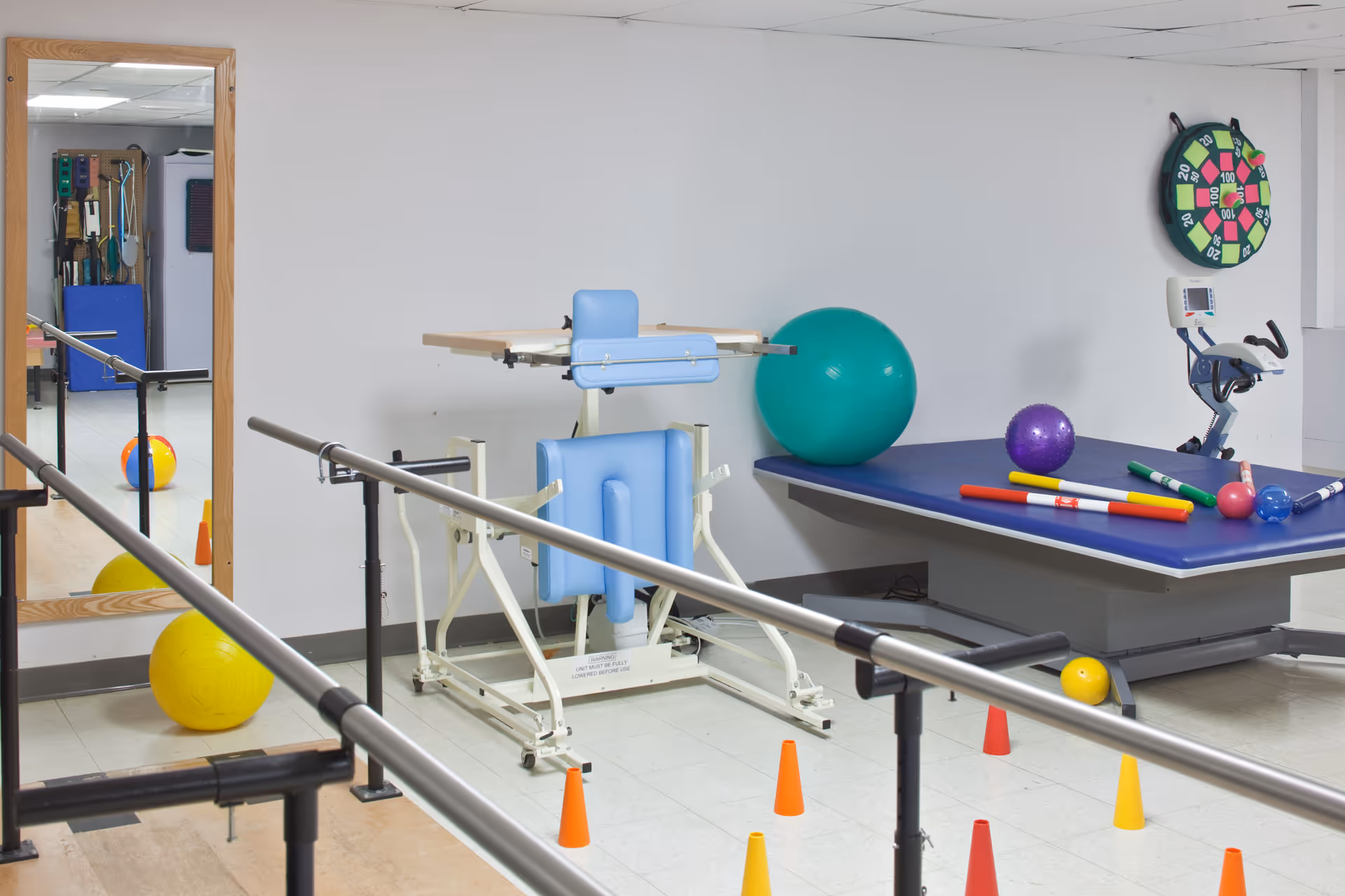 Physical therapy room with parallel bars for walking exercises, a padded exercise table with various therapy balls and colorful rods, a stationary exercise bike, a large mirror reflecting more therapy equipment, and small orange and yellow cones on the floor.