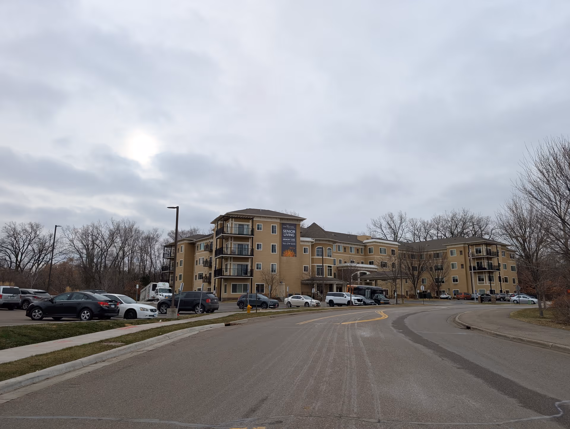 Exterior view of a large senior living facility building with multiple floors and balconies. Several cars are parked in front of the building along a curved driveway. Leafless trees and a cloudy sky are visible in the background.