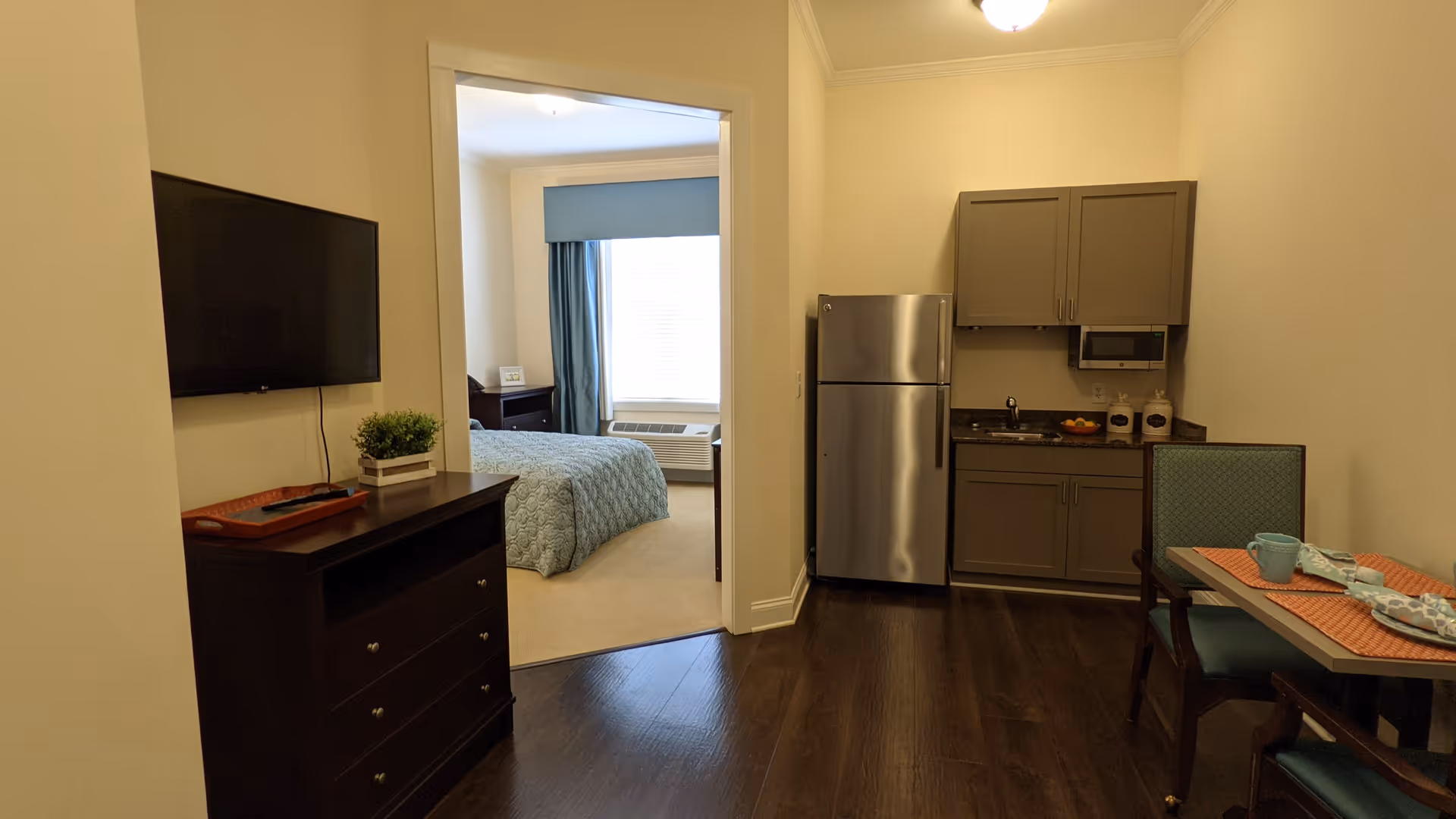 View of a small apartment interior showing a kitchenette with a refrigerator, microwave, and cabinets on the right. A small dining table with two chairs is set with placemats, napkins, and cups. To the left, there is a dresser with a flat-screen TV mounted above it. Through an open doorway, a bedroom with a bed covered in a blue quilt and a window with blue curtains is visible.