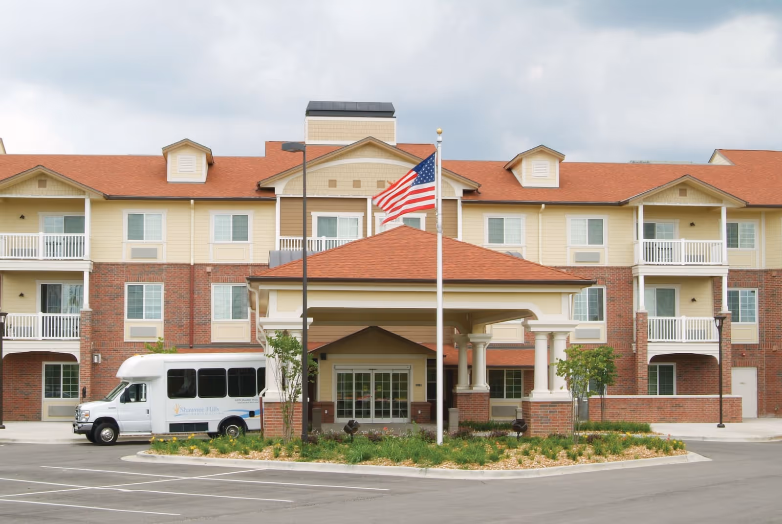 Exterior view of a three-story senior living facility with a covered entrance, an American flag on a flagpole, and a white shuttle bus parked nearby. The building has a combination of brick and beige siding with red roofing and balconies.
