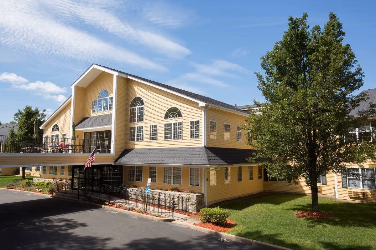 Front exterior of a large yellow two-story senior living building with a balcony, entrance canopy, American flag, and landscaped lawn.