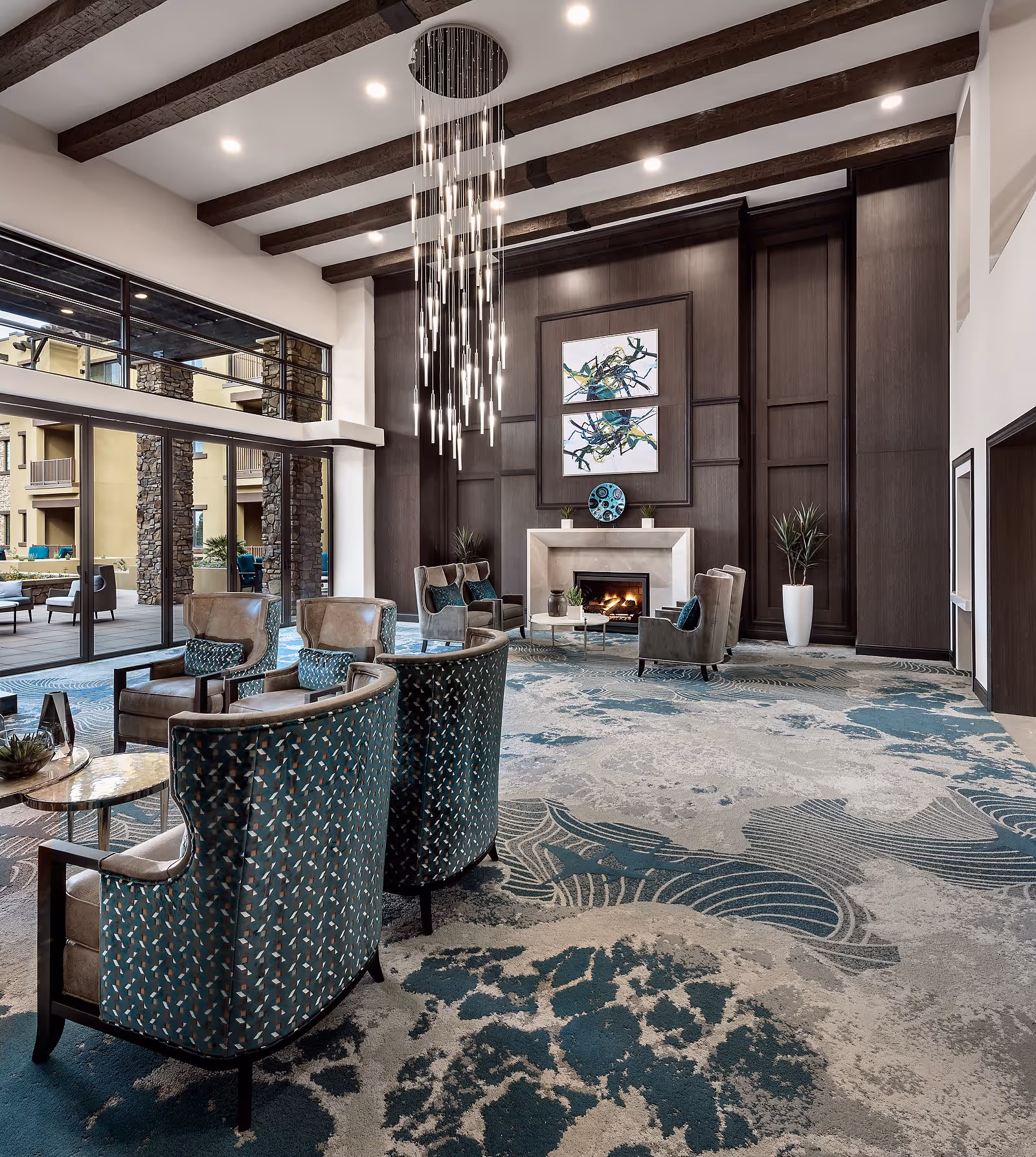A spacious and elegant living room area with high ceilings featuring exposed wooden beams and a modern chandelier. The room has a large patterned carpet in shades of blue and beige, several upholstered armchairs arranged around small tables, a fireplace with decorative art above it, and large windows and glass doors opening to an outdoor patio area.