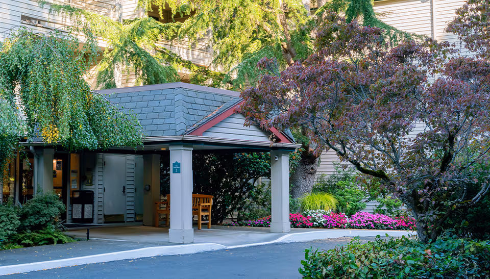 Entrance area of a senior living facility with a covered drop-off zone supported by columns, surrounded by lush greenery including trees, bushes, and colorful flowers. The building exterior is visible in the background.