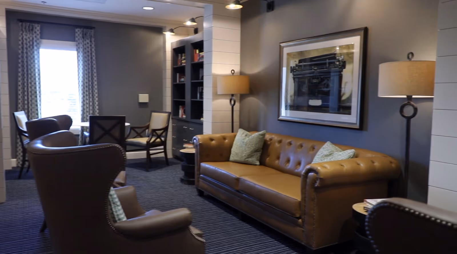 Comfortable furnished lounge with a tufted leather sofa, armchairs, floor lamps, bookshelf, and a small table by a window.