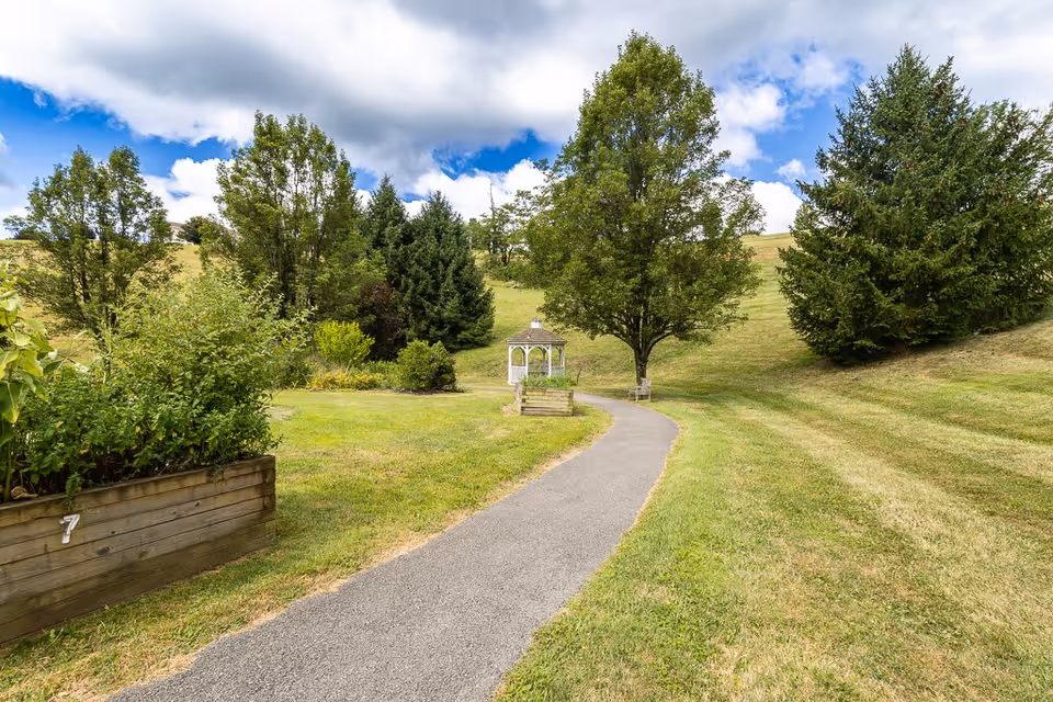 A paved walking path winds through a grassy outdoor area with various trees and shrubs. There is a small white gazebo along the path and a wooden raised garden bed marked with the number 7. The sky is partly cloudy with patches of blue visible.
