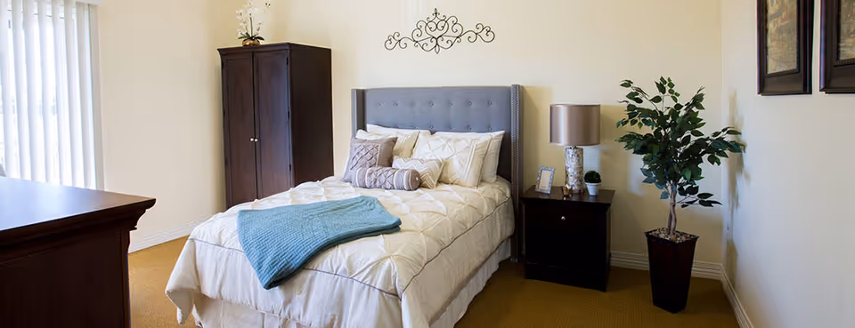 A neatly made bed with a gray upholstered headboard, white bedding, and decorative pillows in a bedroom. A blue throw blanket is draped over the bed. To the right of the bed is a dark wooden nightstand with a lamp, a small plant, and a framed photo. A tall potted plant is placed next to the nightstand. On the left side of the room, there is a dark wooden dresser and a tall dark wooden cabinet. The walls are light-colored, and there is a decorative wall hanging above the bed. Vertical blinds cover a window on the left side.