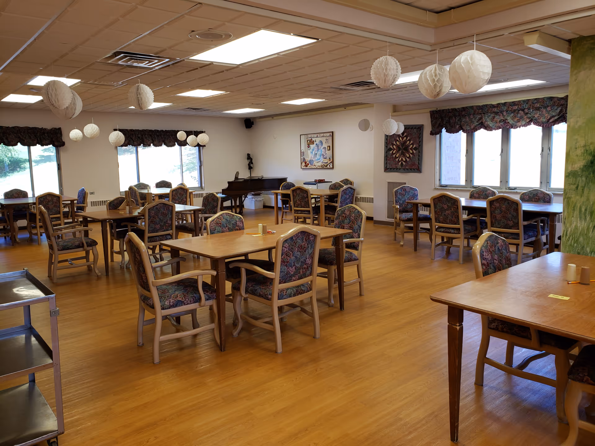 Spacious senior living dining room with multiple wooden tables and floral-upholstered chairs, paper lantern decorations, and large windows.