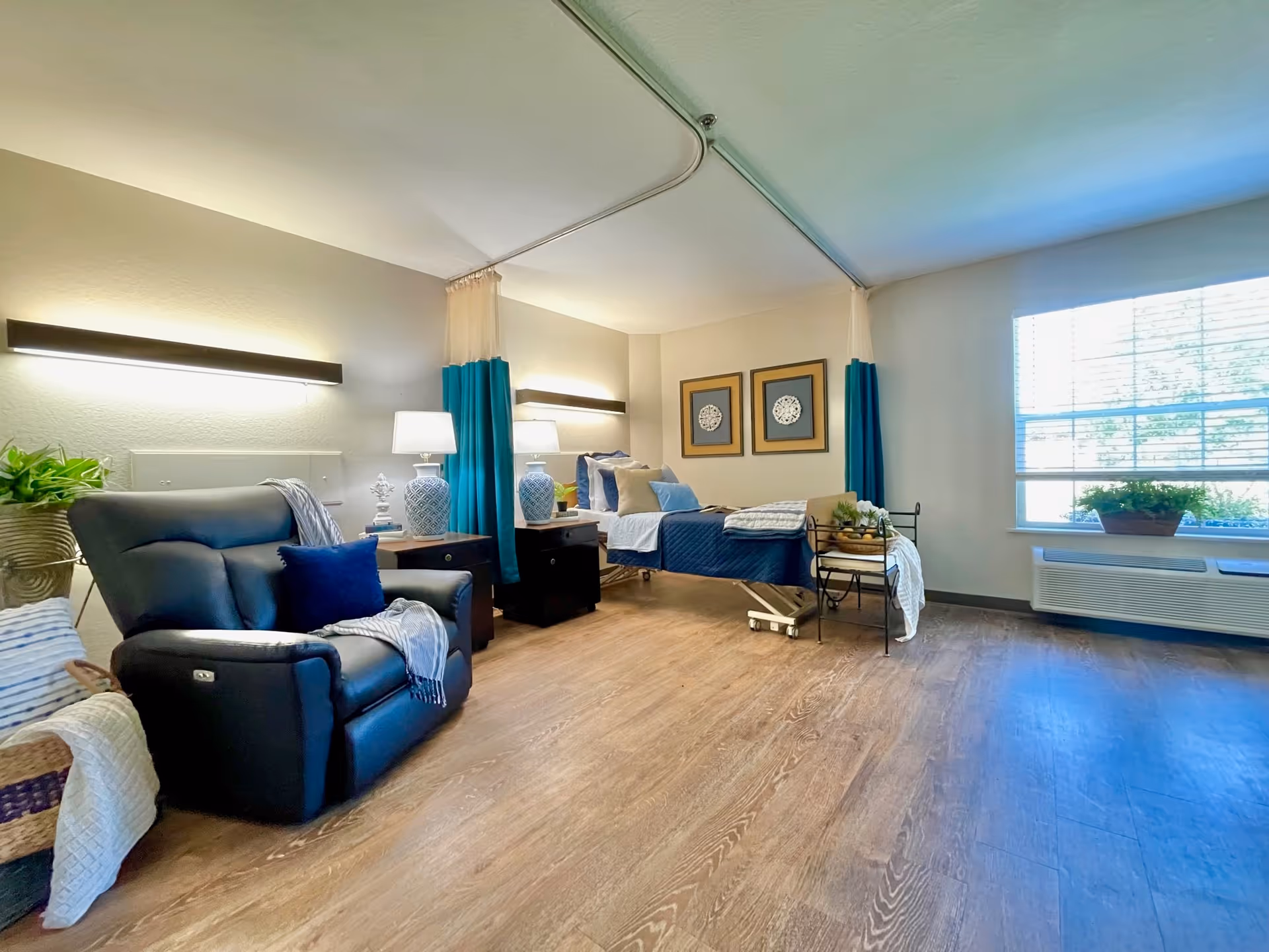 A spacious and well-lit bedroom in a senior living facility featuring a single bed with blue and white bedding, two bedside tables with lamps, a comfortable black recliner chair with a blue pillow and a throw blanket, framed artwork on the wall, large window with blinds, and wooden flooring.