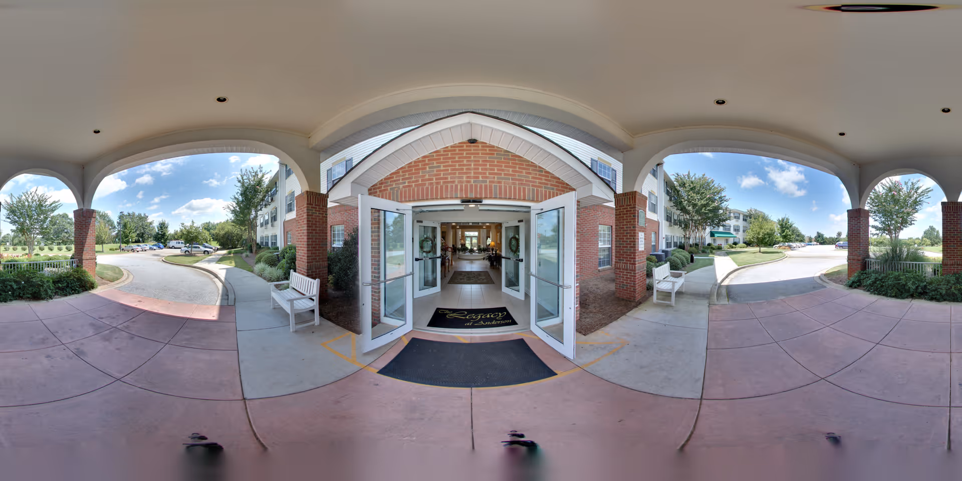 Covered entrance area of The Legacy of Anderson facility with open double glass doors leading inside. The entrance is framed by brick pillars and white benches on either side. Outside, there is a curved driveway with landscaped greenery and trees under a blue sky with scattered clouds.