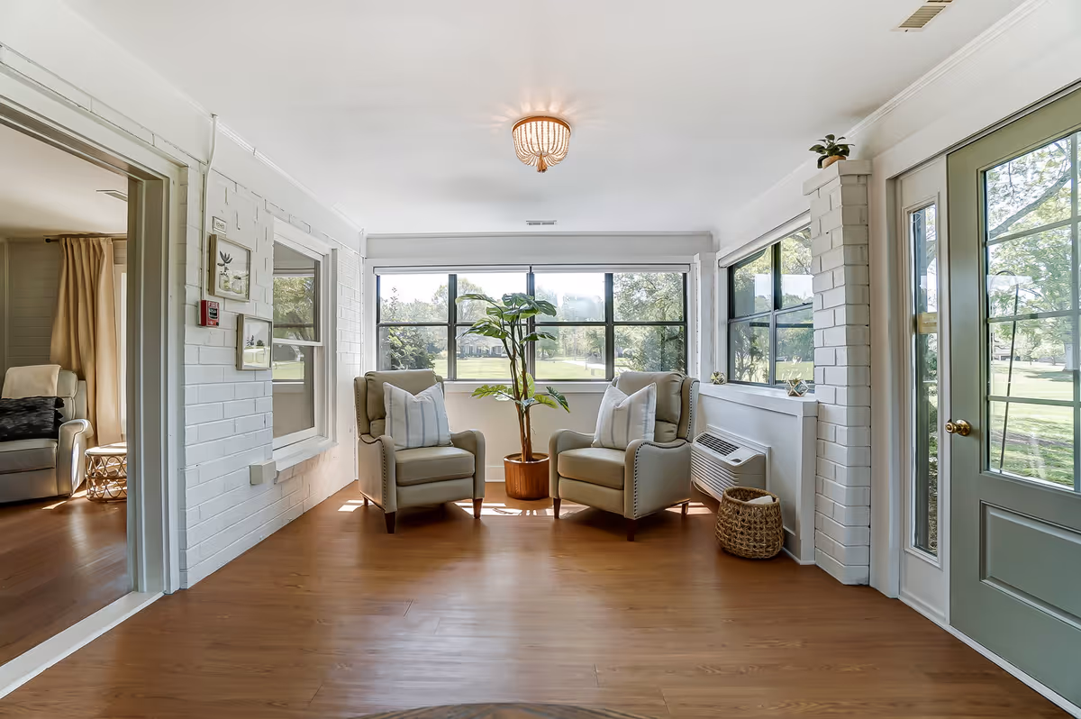 A bright sitting area with two beige armchairs with white pillows facing each other, a potted plant between them, large windows showing a green outdoor view, and a green door with glass panels on the right. The room has white brick walls and wooden flooring.