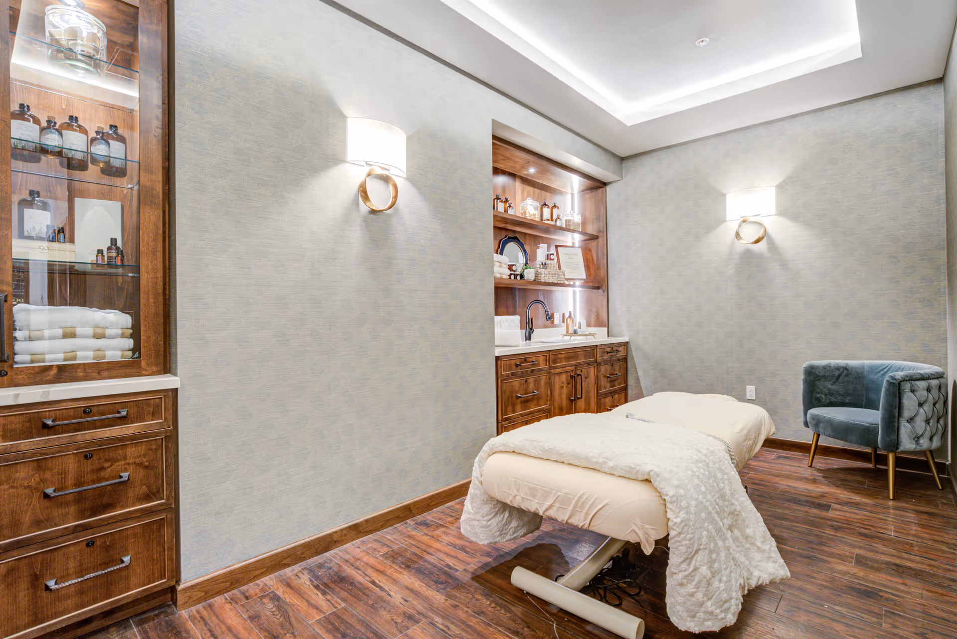 A serene treatment room with a massage table covered in a white blanket, wooden cabinets with glass shelves displaying bottles and towels, soft wall sconces, a blue upholstered chair, and warm wooden flooring.