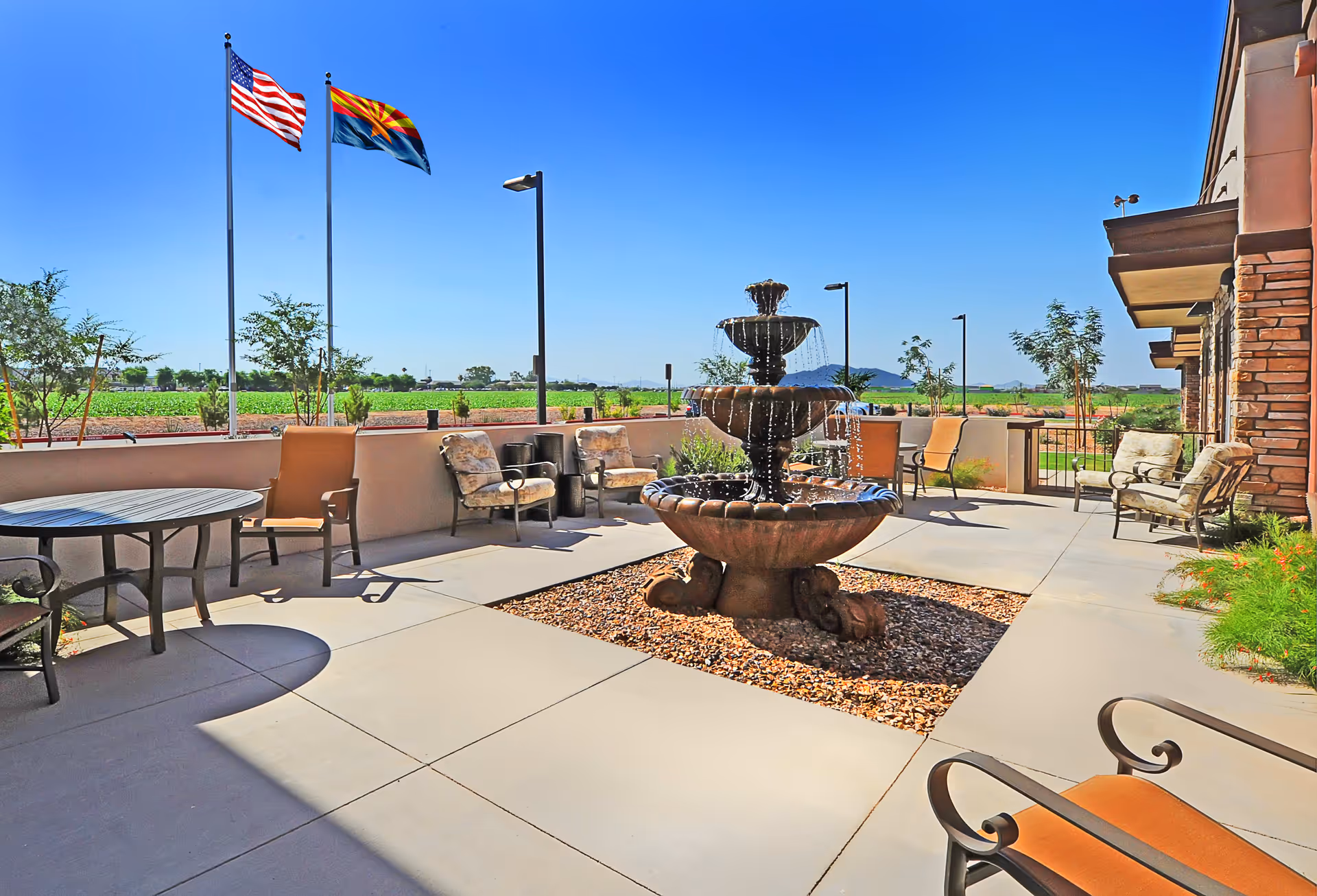 Outdoor patio area at Sky Ridge Senior Living featuring a three-tiered water fountain in the center, surrounded by cushioned chairs and tables. Two flagpoles display the American flag and the Arizona state flag against a clear blue sky. The patio is adjacent to a building with stone and stucco exterior walls, and there are landscaped plants and trees around the seating area.