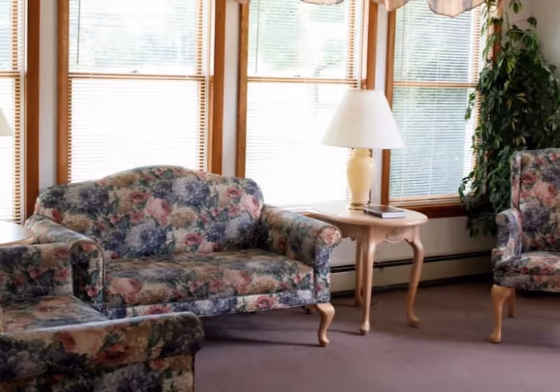 A cozy living room area with floral patterned upholstered sofa and armchairs, a wooden side table with a cream-colored lamp and a book, large windows with blinds, and a tall green plant in the corner.
