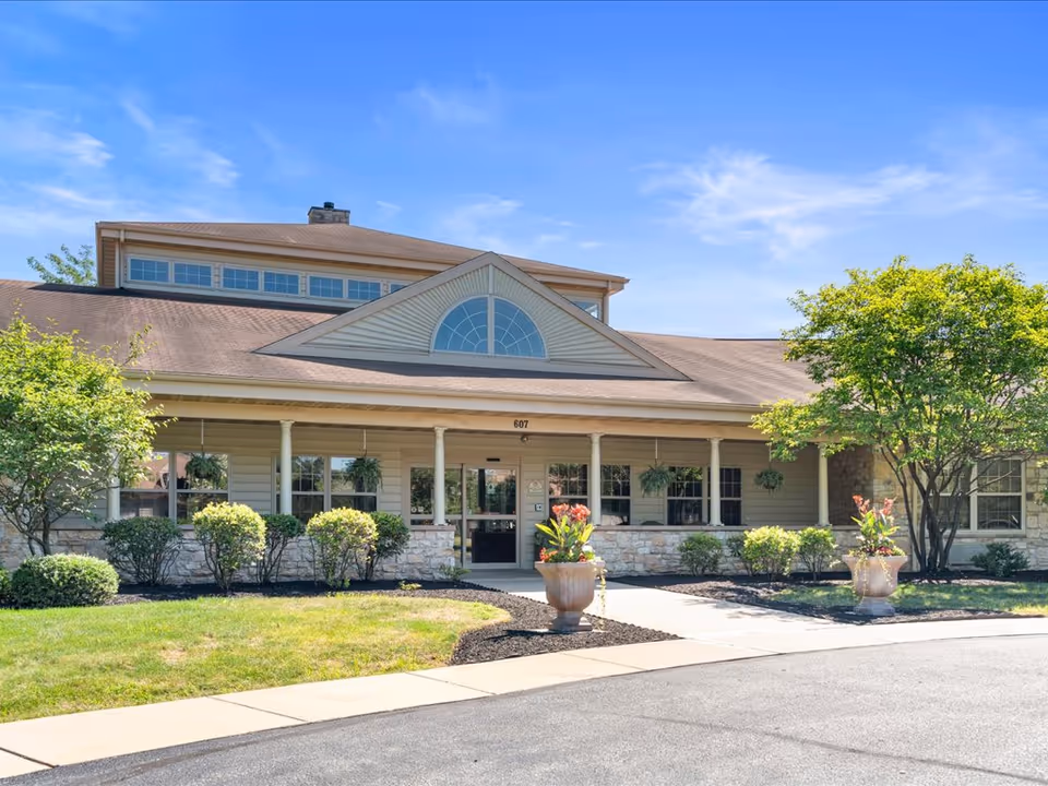 Front exterior view of a single-story building with a peaked roof and large windows. The entrance has a covered porch supported by columns, with two large flower pots containing plants on either side of the walkway. There are shrubs and small trees around the building, and the sky is clear and blue.