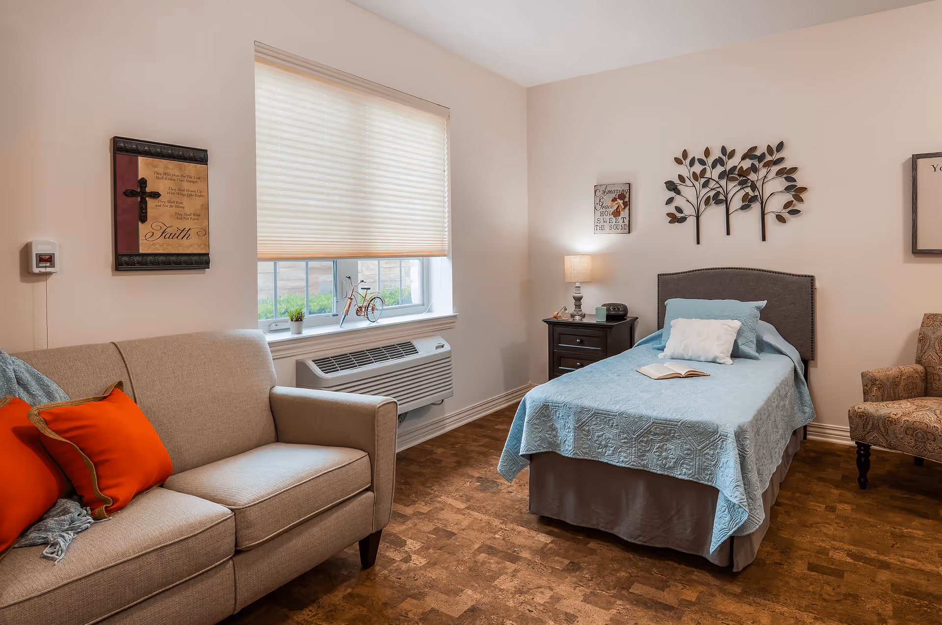 A cozy bedroom in a senior living facility featuring a single bed with a light blue quilt and pillows, a nightstand with a lamp and clock, a beige sofa with red cushions, a patterned armchair, a window with blinds, and decorative wall art including a metal tree sculpture and framed inspirational quotes.