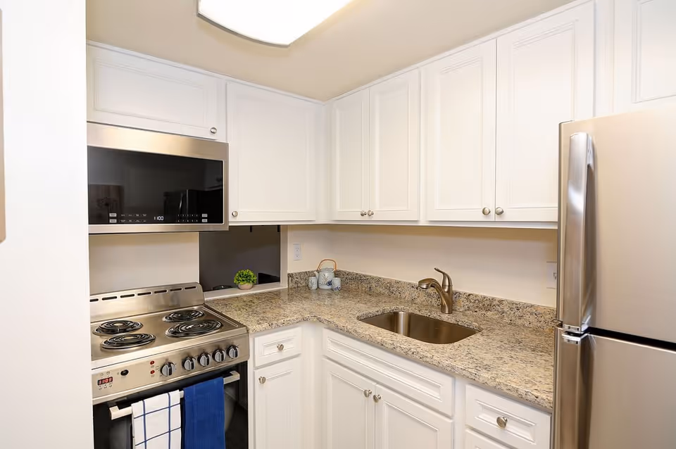 A compact kitchen with white cabinets, a granite countertop, a stainless steel electric stove with four coil burners, a built-in microwave above the stove, a stainless steel sink with a bronze faucet, and a stainless steel refrigerator. A blue and white towel hangs on the oven handle, and a small green plant and decorative items are on the countertop.