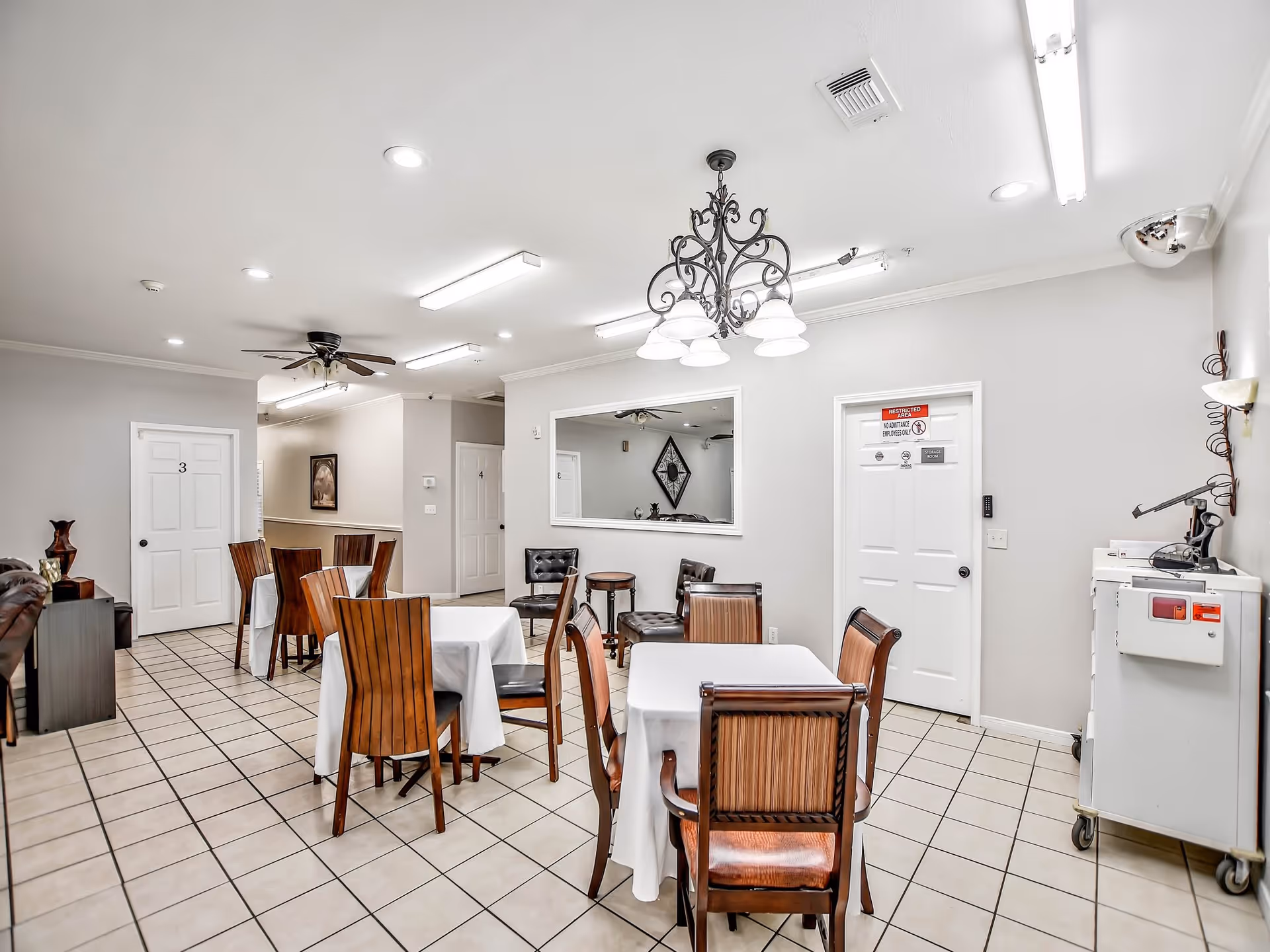 Interior view of a dining area in an assisted living facility with several tables covered with white tablecloths and wooden chairs around them. The room has tiled flooring, white walls, and multiple doors labeled with numbers and signs. A decorative chandelier hangs from the ceiling, and there is a large mirror on one wall reflecting part of the room. Additional seating with black chairs and a small round table is visible in the background.