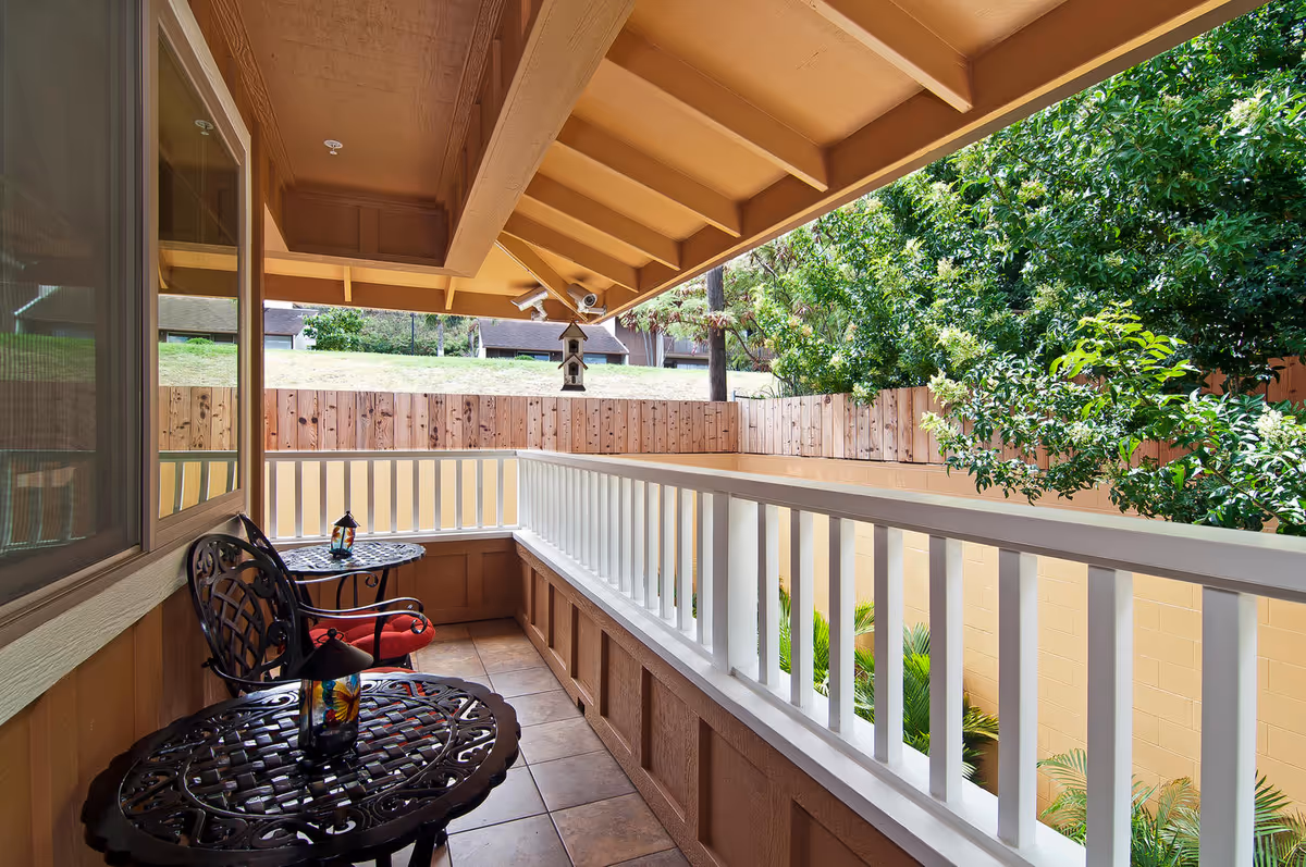 A covered outdoor balcony area with two black metal chairs with red cushions and two matching round tables, each holding a decorative lantern. The balcony has a white railing and overlooks a fenced yard with green trees and shrubs.