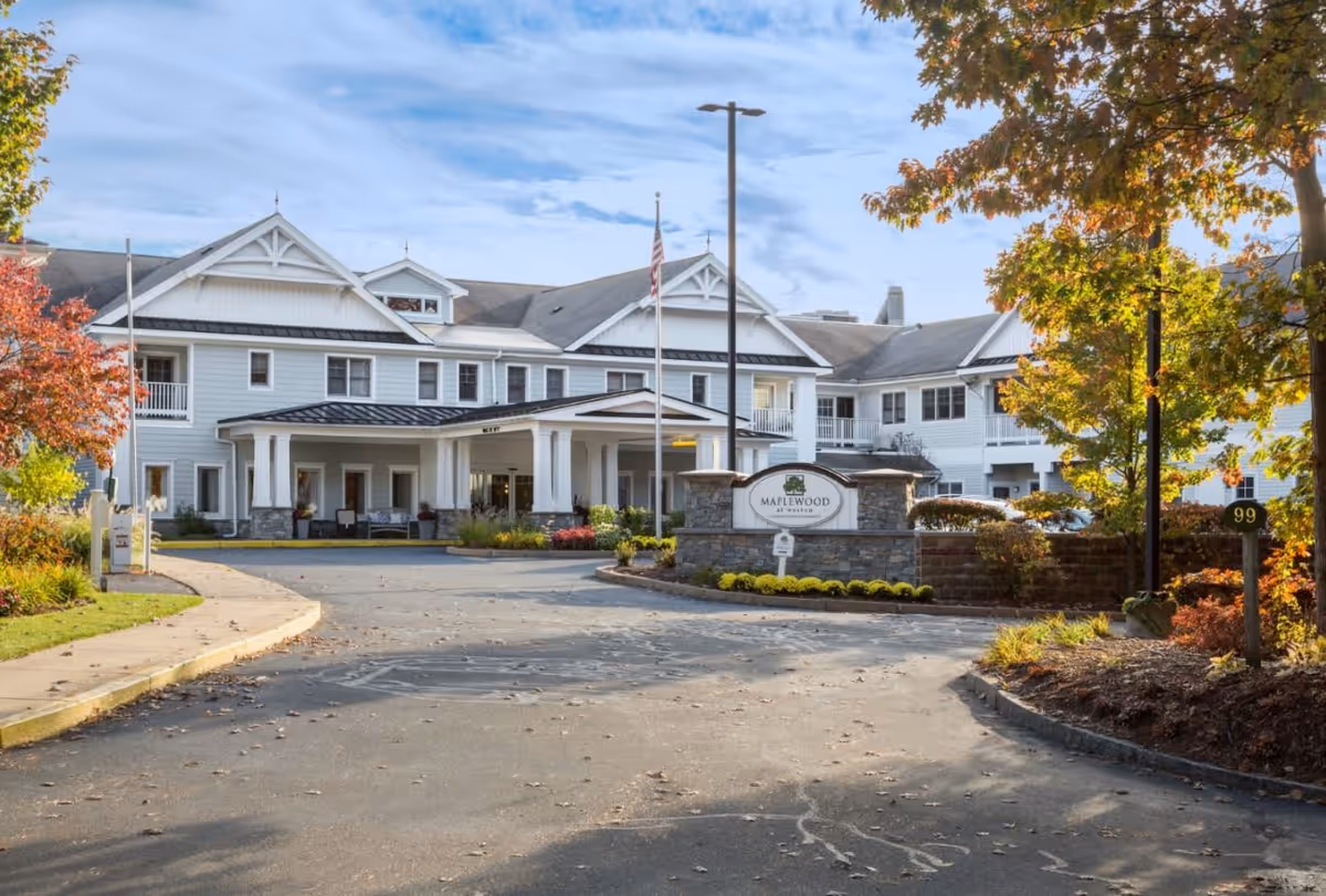 Front exterior view of Maplewood at Weston, a large two-story white building with a covered entrance, surrounded by trees with autumn foliage and a driveway leading up to the entrance.