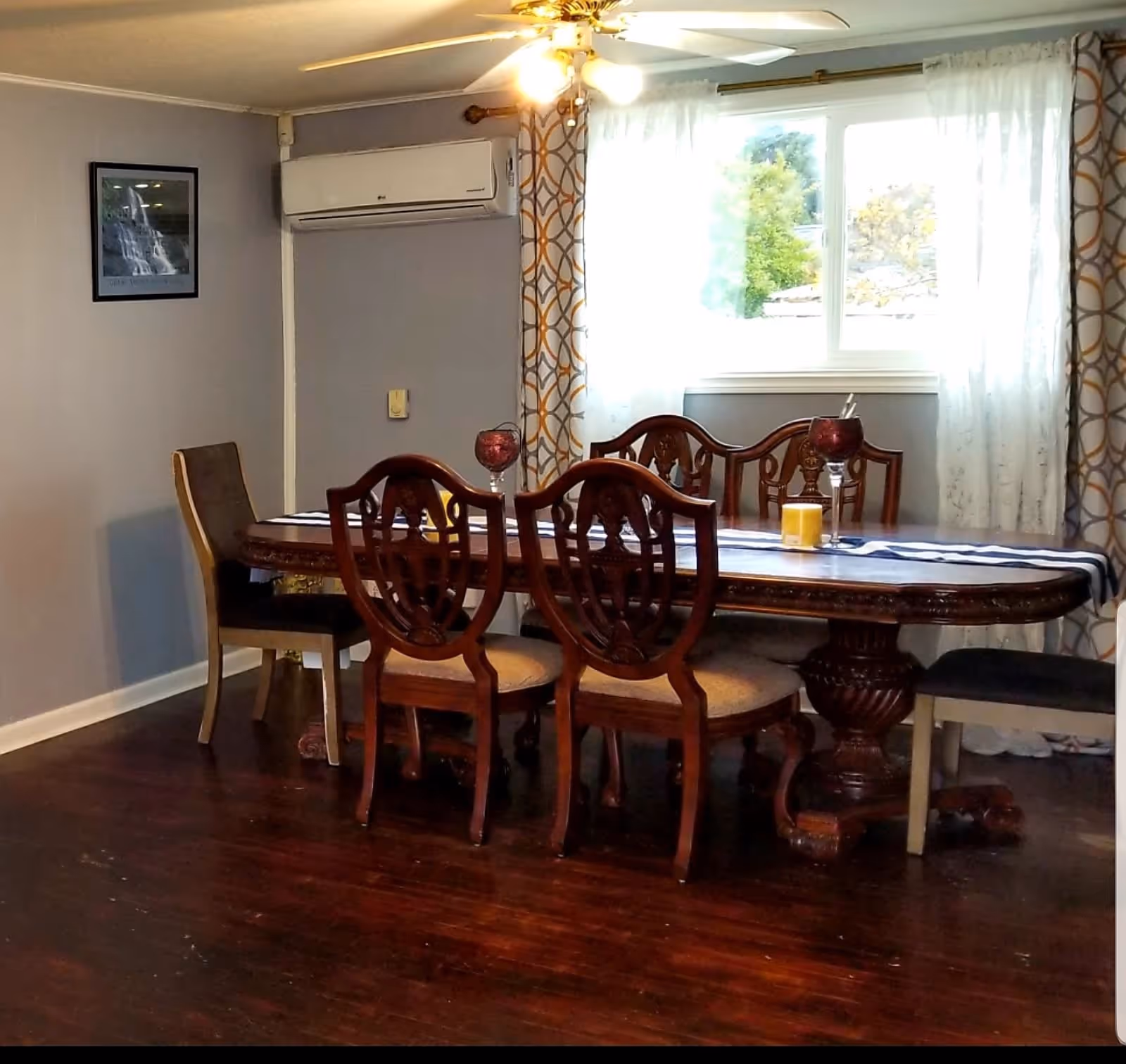 Ornate wooden dining table with chairs under a ceiling fan in a bright room with a window and patterned curtains.