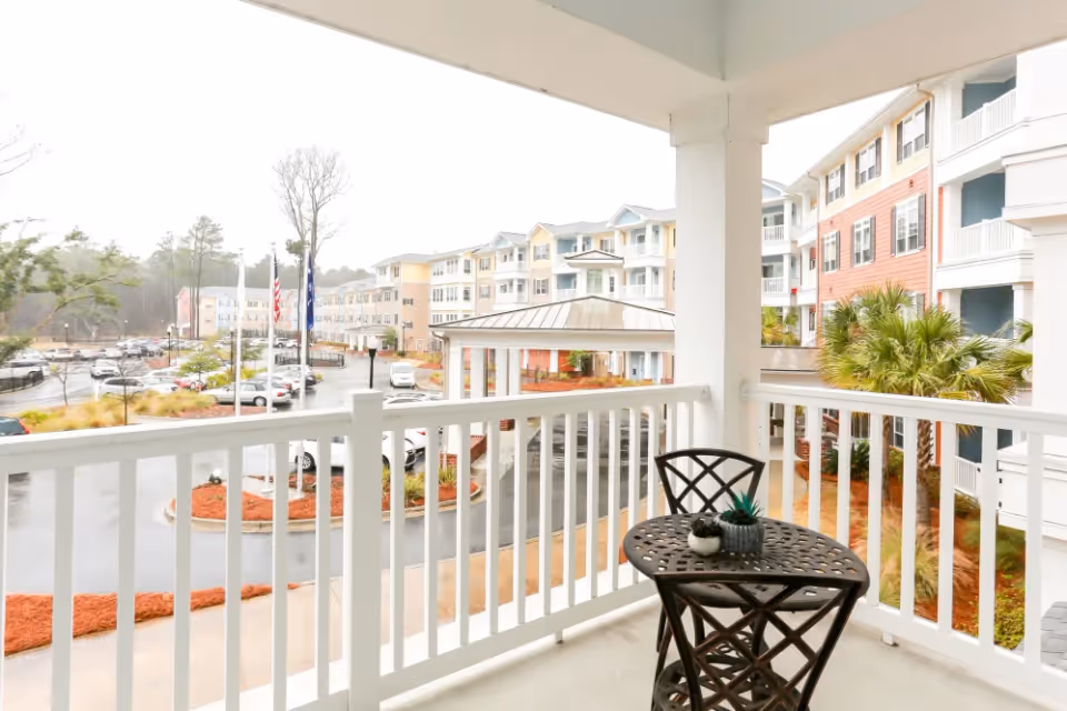 View from a covered balcony with a small round metal table and two matching chairs. The balcony overlooks a parking lot, landscaped areas with mulch and small plants, and a multi-story residential building with balconies and a covered entrance.