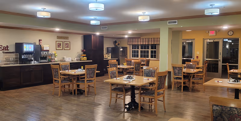 A well-lit dining room in a senior living facility with several wooden tables and chairs arranged neatly. The room features a beverage station with a soda fountain and coffee maker against one wall, decorative framed pictures, and a window with plaid curtains. The floor is wooden, and ceiling lights provide warm illumination.