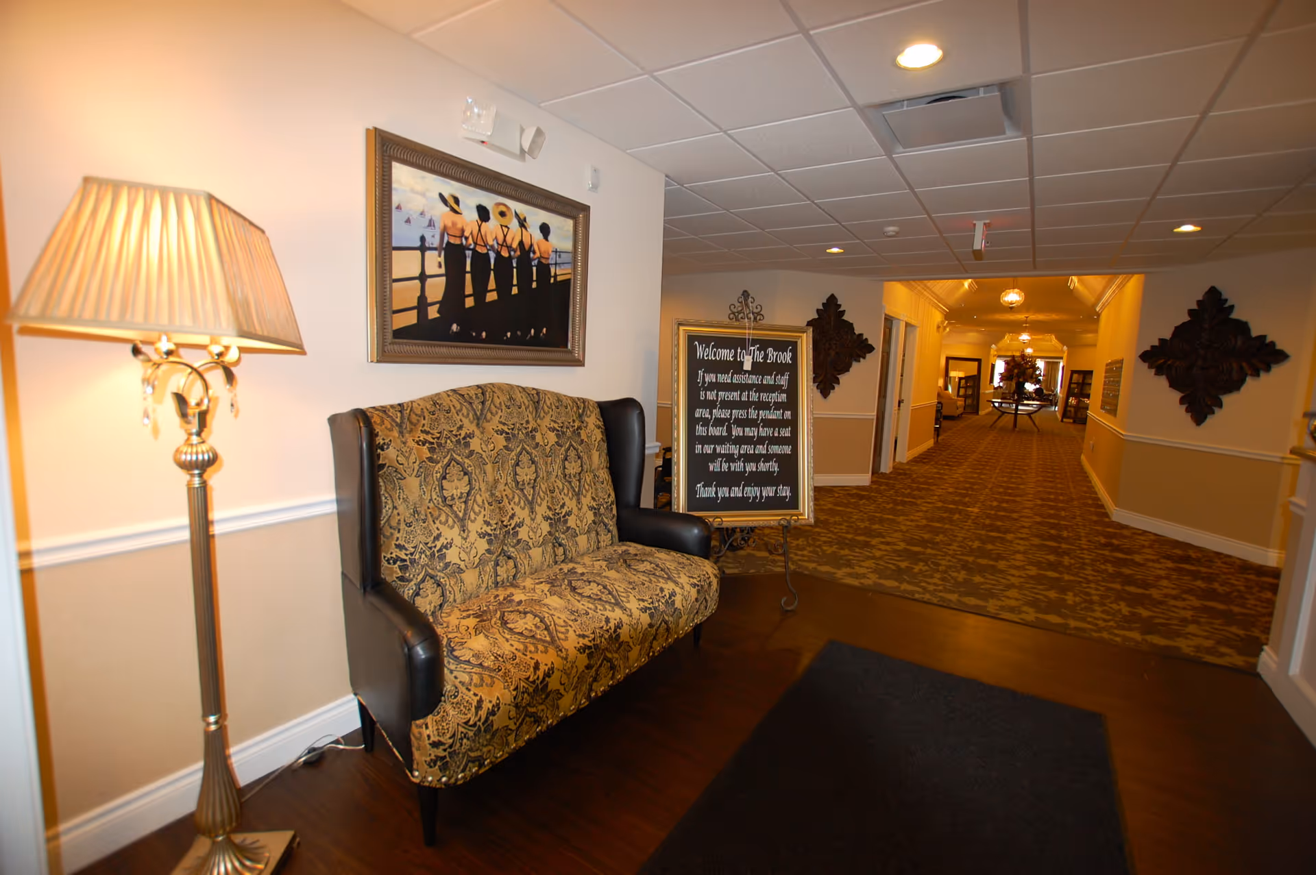 A hallway in The Brook of Boyne City featuring a patterned loveseat with black armrests, a standing lamp with a pleated shade, a framed painting of five women in black dresses and hats looking at sailboats, and a sign welcoming visitors with instructions for assistance. The hallway has carpeted flooring with a floral pattern, beige walls with white trim, and decorative wall hangings.