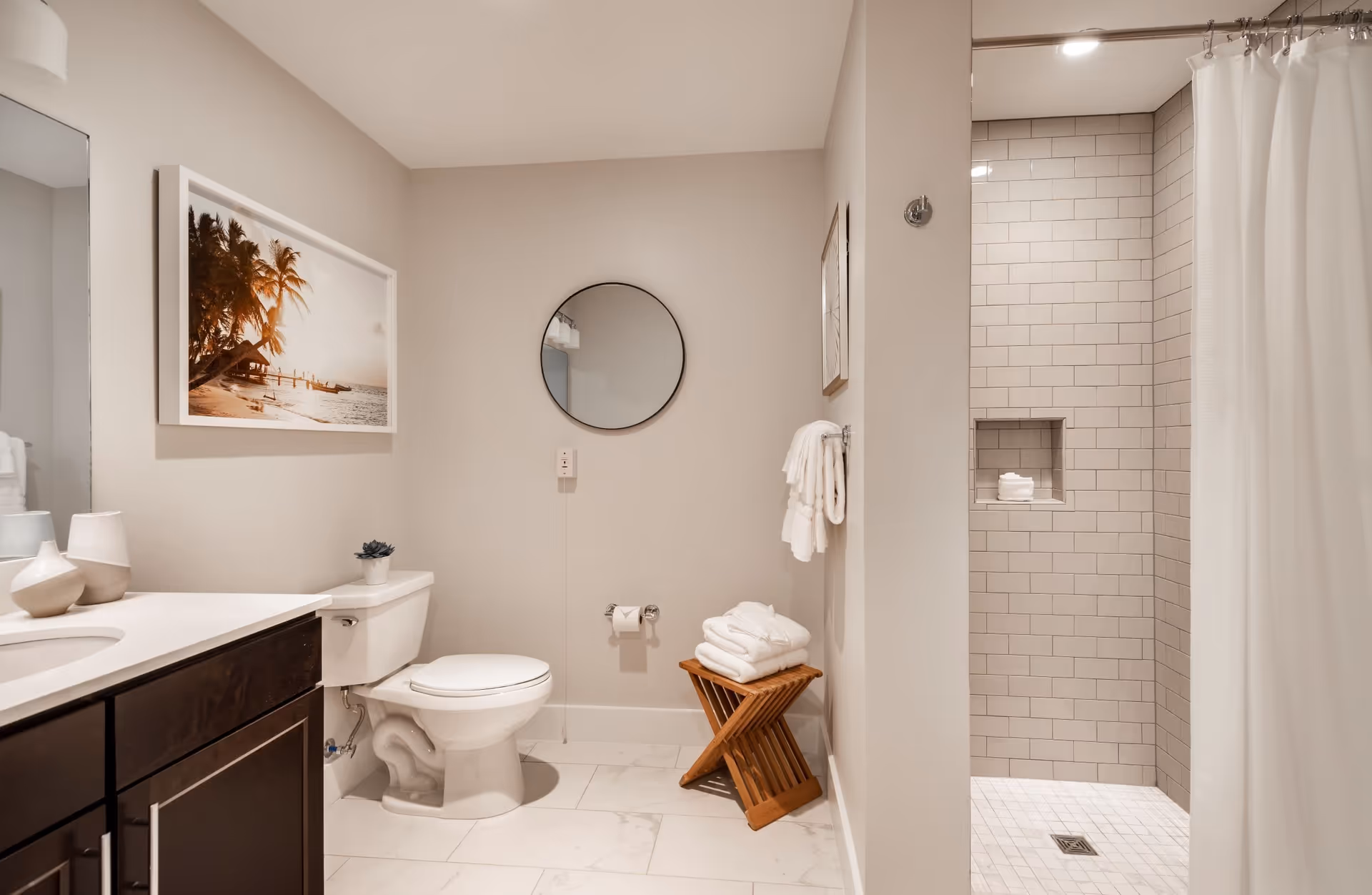 A modern bathroom with a white toilet, dark wood vanity with a white countertop, a round mirror on the wall, a framed picture of a beach scene, a wooden stool with folded white towels, and a tiled walk-in shower with a white curtain.