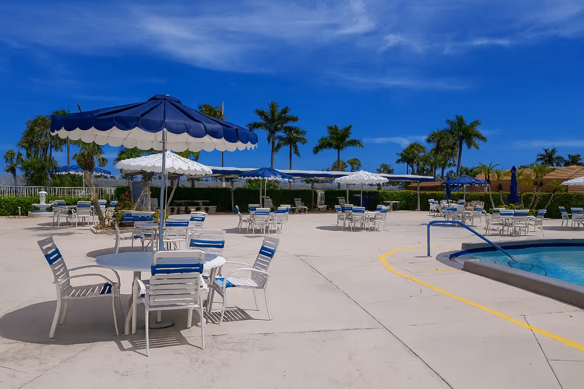 Outdoor poolside patio with tables, chairs, blue-and-white umbrellas and palm trees under a clear blue sky.