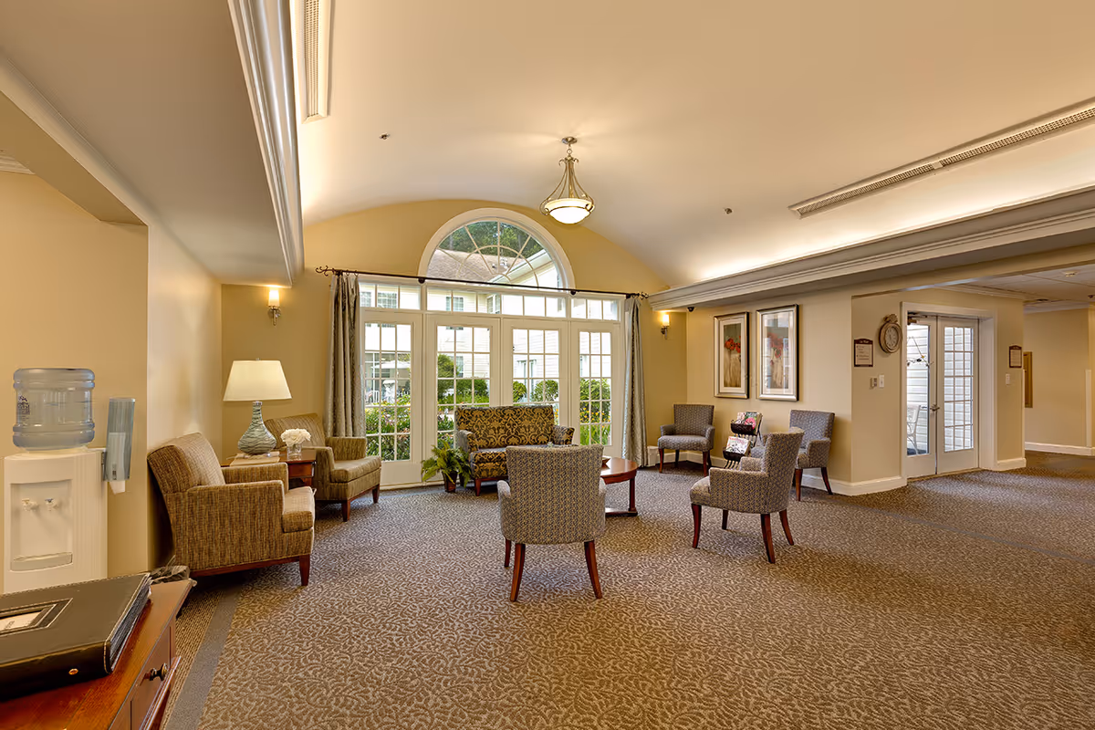 A spacious and well-lit common area in a senior living facility with several upholstered chairs and a loveseat arranged around a wooden coffee table. Large windows with an arched top allow natural light to fill the room, and there are framed artworks on the walls. A water cooler and a lamp on a side table are visible on the left side, and double glass doors lead to another area on the right.
