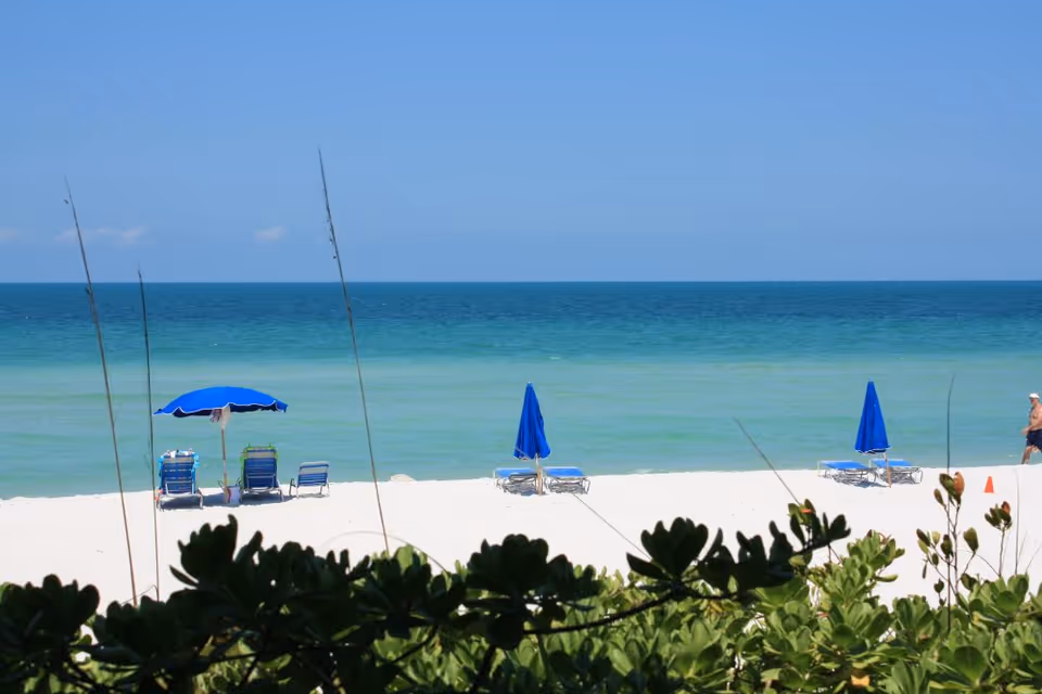 A serene beach scene with white sand, blue ocean water, and a clear blue sky. There are several blue beach umbrellas and lounge chairs set up on the sand. Green foliage is visible in the foreground, and a person is walking along the beach on the right side of the image.