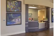 Reception area of a nursing and rehabilitation center with a curved front desk where a receptionist is seated. The walls are decorated with two framed paintings depicting nature scenes. The floor has a wood-like finish and the lighting is bright with ceiling fixtures.