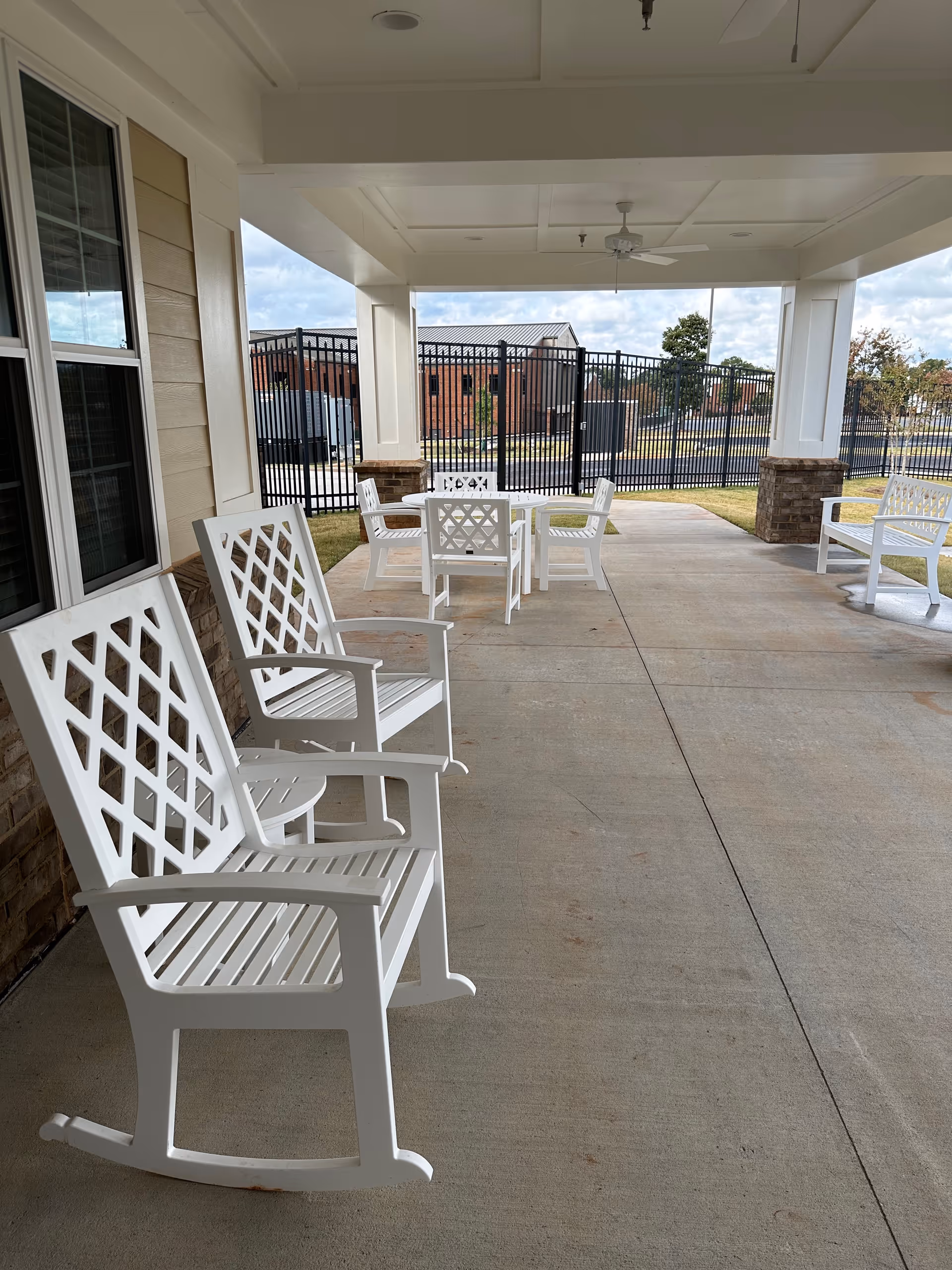 Covered outdoor patio with white rocking chairs and tables on a concrete floor overlooking a fenced lawn.