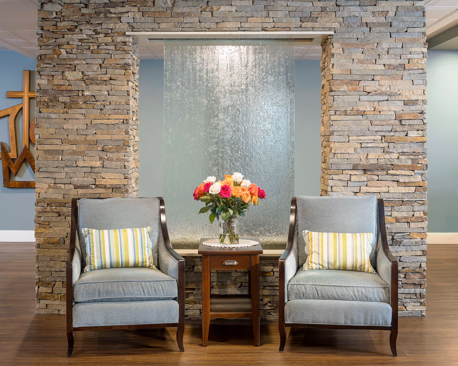 Two light blue upholstered armchairs with striped cushions are positioned on either side of a small wooden table holding a vase of colorful roses. Behind the table is a textured glass water feature set within a stone wall. The floor is wooden, and a wooden decorative wall art piece is partially visible on the left.