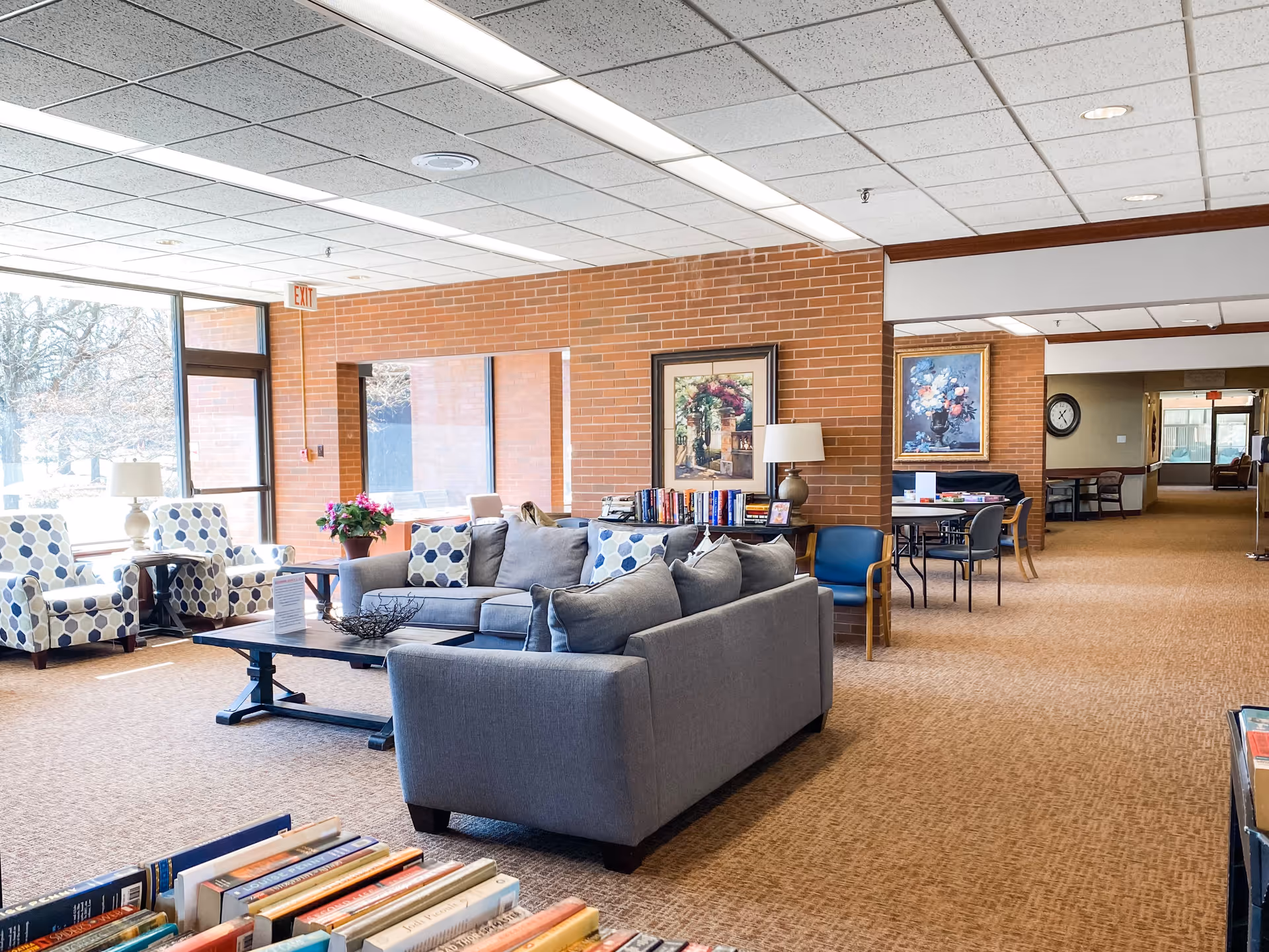 A bright and spacious senior living facility common area with large windows letting in natural light. The room features a gray sectional sofa with patterned pillows, two armchairs with blue and white geometric patterns, a coffee table, and a side table with a lamp. There are framed paintings on the brick wall, a bookshelf with books, and a table with chairs in the background. The carpet is beige, and the ceiling has white tiles with fluorescent lighting.