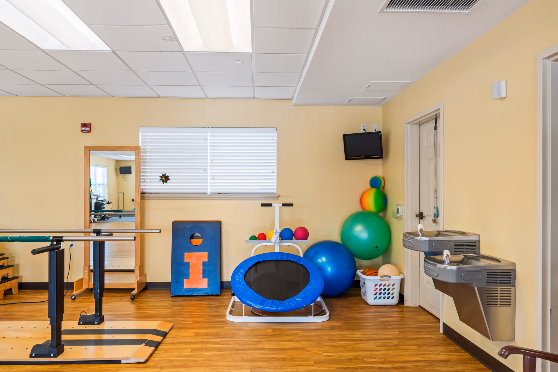 An interior therapy/exercise room with parallel bars, a small trampoline, colorful exercise balls, and wall-mounted water fountains.