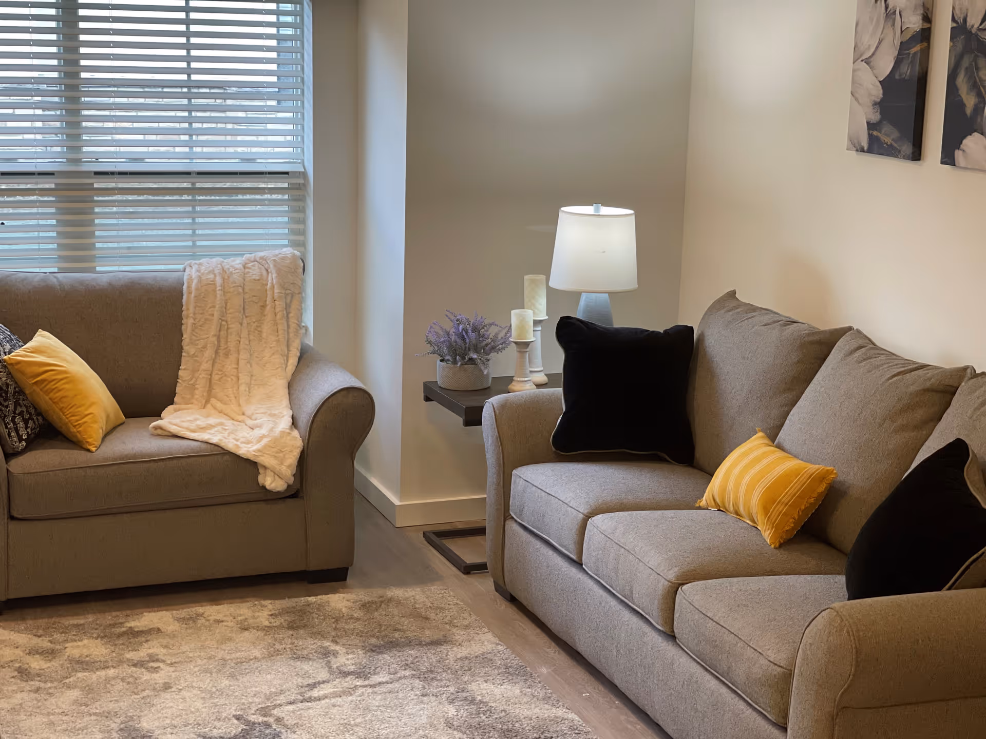A cozy living room with two gray sofas adorned with black and yellow pillows. A soft white throw blanket is draped over one sofa. Between the sofas is a small side table holding a lamp, two candles, and a small potted plant. The room has a large window with white blinds and light-colored walls with floral artwork.