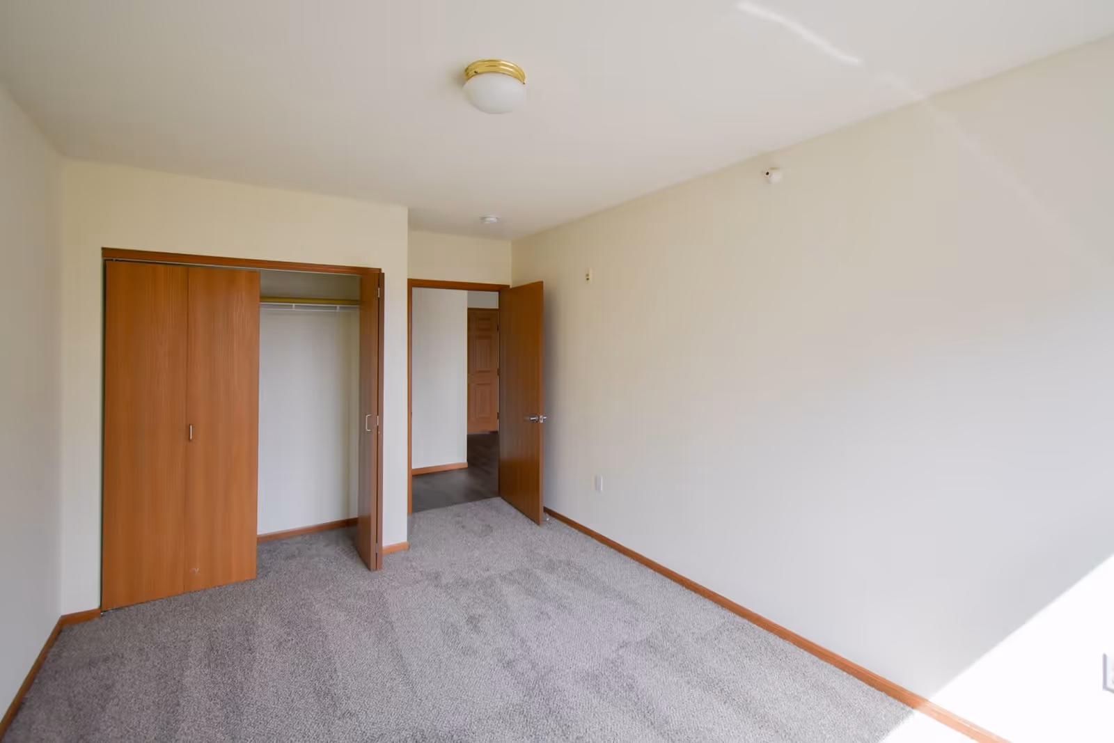 Empty bedroom with beige walls, gray carpet, a wooden closet with sliding doors partially open, and a wooden door leading to another room. The room is well-lit with natural light coming from the right side.