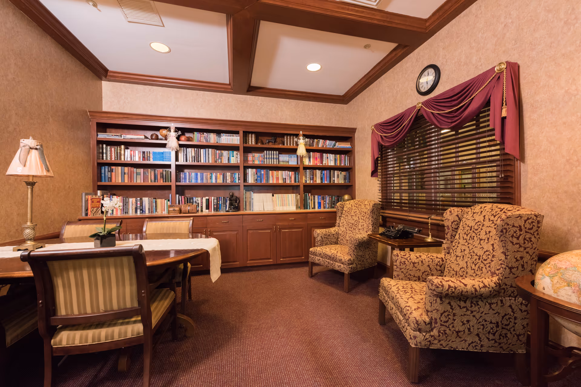 A cozy library or reading room with a large wooden bookshelf filled with books, two patterned armchairs, a wooden table with chairs, a lamp, and a globe. The room has warm beige walls, a carpeted floor, and a window with wooden blinds and a burgundy valance with gold tassels. A clock is mounted above the window.