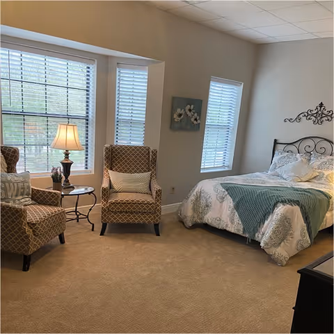 A cozy bedroom with a bed featuring a decorative metal headboard, white and green bedding, and a green throw blanket. Two patterned armchairs with pillows are positioned near two windows with blinds. A small round table with a lamp and a plant sits between the chairs. The walls are light-colored with a floral wall decoration above the bed.