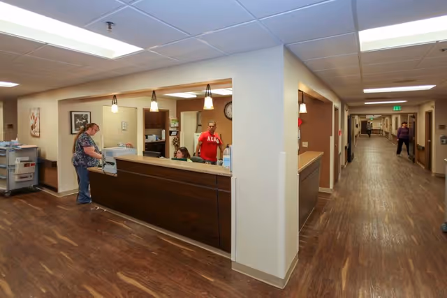 Interior view of a healthcare facility reception area with a wooden front desk, three people behind and near the desk, and a long hallway extending to the right with two people walking. The floor is wood-patterned, and the ceiling has recessed lighting and hanging pendant lights above the desk.