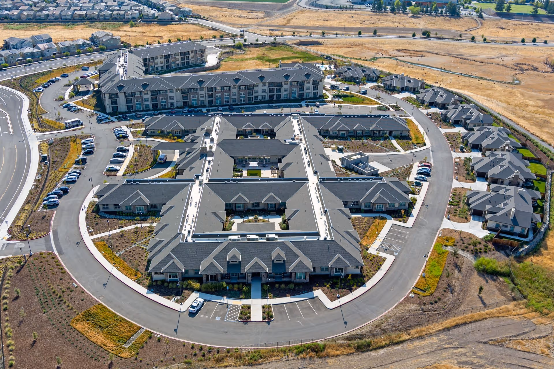 Aerial view of Ansel Park Independent Living facility showing multiple connected buildings with gray roofs arranged in a circular layout, surrounded by parking lots, roads, and landscaped areas. The surrounding area includes dry grassland and residential neighborhoods in the distance.